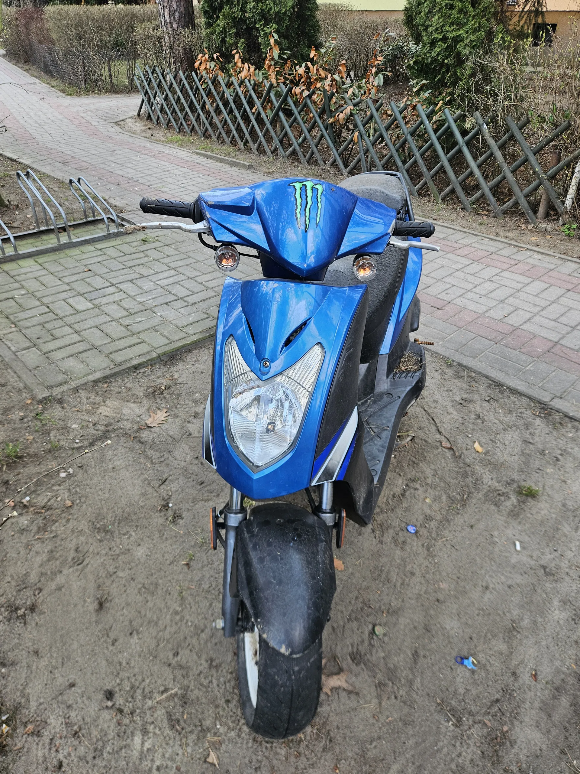 Niebieski skuter z zielonym logo Monster Energy zaparkowany na brudnej ziemi obok chodnika z czerwono-szarymi płytkami. W tle metalowe ogrodzenie, krzewy z suchymi liśćmi i fragment budynku. Skuter ma czarną maskę przednią i reflektor. Na ziemi widoczne są rozrzucone liście i drobne śmieci.