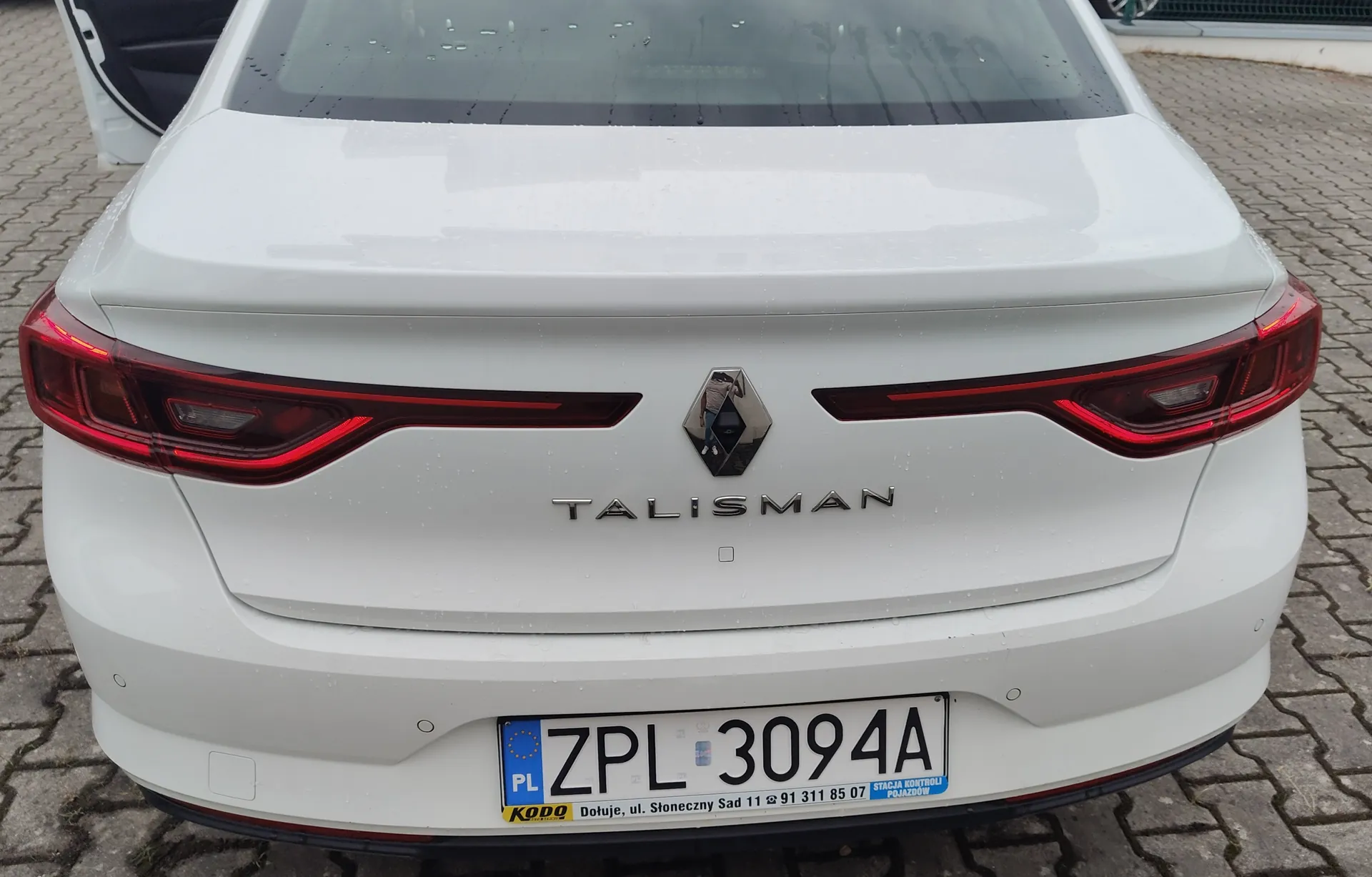 Tylny widok białego samochodu marki Renault Talisman. Na klapie bagażnika znajduje się logo Renault oraz napis 'TALISMAN'. Samochód posiada polską tablicę rejestracyjną o numerze ZPL 3094A. Poniżej tablicy widnieje napis 'KODO' oraz adres i numer telefonu: 'ul. Słoneczny Sad 11 91 311 85 07'. Auto zaparkowane jest na kostce brukowej.
