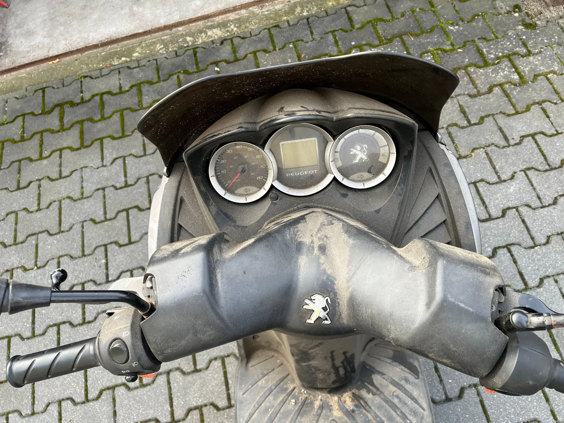 Widok z perspektywy kierowcy na kierownicę i deskę rozdzielczą zakurzonego skutera marki Peugeot. Na desce znajdują się dwa analogowe wskaźniki oraz centralny wyświetlacz z napisem PEUGEOT. Na obudowie kierownicy widoczne jest srebrne logo lwa. Skuter stoi na kostce brukowej.
