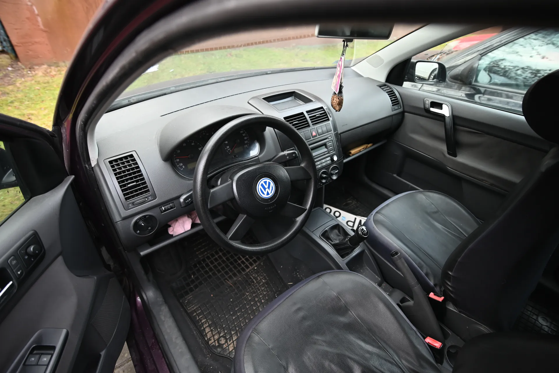 Wnętrze samochodu marki Volkswagen z widocznym kierownicą z logo VW, czarnymi skórzonymi fotelami, szarą deską rozdzielczą z wentylacjami i radioodtwarzaczem. Drzwi po stronie kierowcy są otwarte, na zewnątrz widać trawnik i ceglaną ścianę. Na lusterku wstecznym wiszą ozdoby. Podłoga pokryta jest czarnymi matami, a na desce rozdzielczej widoczne są wskaźniki prędkości i obrotomierz.
