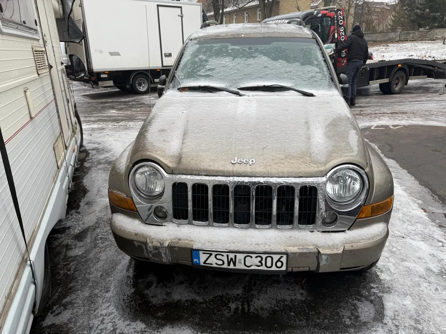 Przedni widok brązowego samochodu Jeep pokrytego śniegiem, z widocznym napisem "Jeep" na masce i tablicą rejestracyjną ZSW C306 z kodem PL. W tle biały przyczepa, ciężarówka z zamkniętym naczepą oraz czerwono-czarny holownik z numerem 6026 022. Na prawo od Jeepsa stoi osoba w ciemnej kurtce i czapce. Tło stanowi zimowy plac z resztkami śniegu na ziemi i budynkami w oddali.