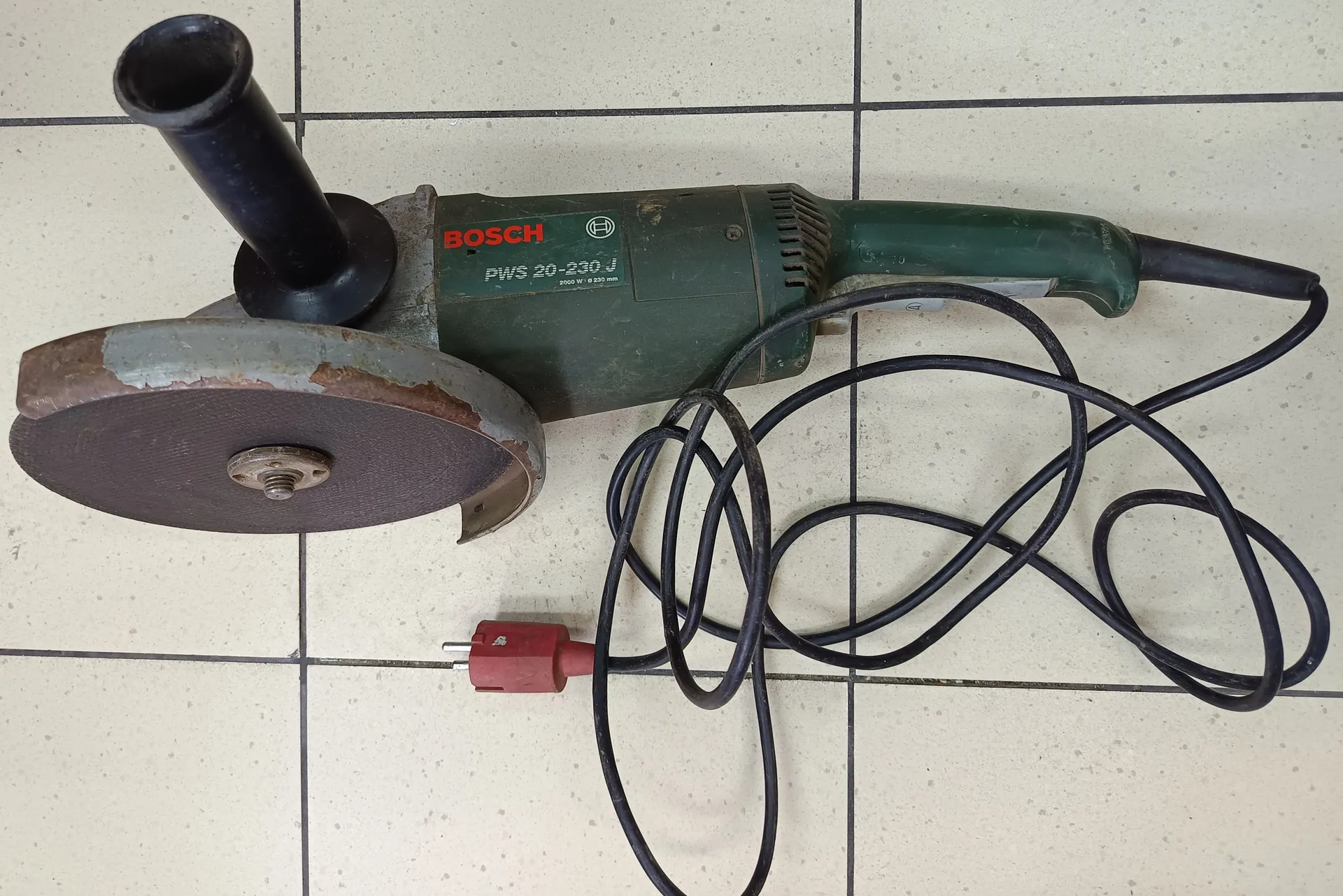 Szlifierka kątowa marki BOSCH model PWS 20-230 J o mocy 2000 W i średnicy dysku 230 mm. Zielone ciało urządzenia z czarną rękojeścią, zużyty dysk z widoczną rdzą, czarny przewód z czerwoną wtyczką leżący na jasnych płytkach podłogowych.