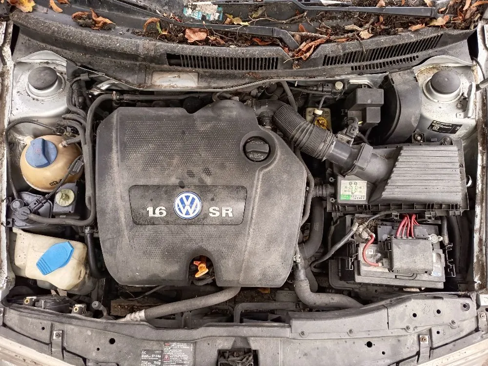 Widok z góry na zakurzoną komorę silnika samochodu. W centrum znajduje się plastikowa osłona silnika z logo Volkswagena oraz napisem '1.6 SR'. Po lewej stronie widoczny jest zbiornik płynu z niebieskim korkiem oraz inny zbiornik z żółtą zakrętką. Po prawej stronie znajduje się akumulator z czerwonymi przewodami oraz obudowa filtra powietrza. Całość jest pokryta warstwą kurzu, a na górnej krawędzi maski leżą suche liście.