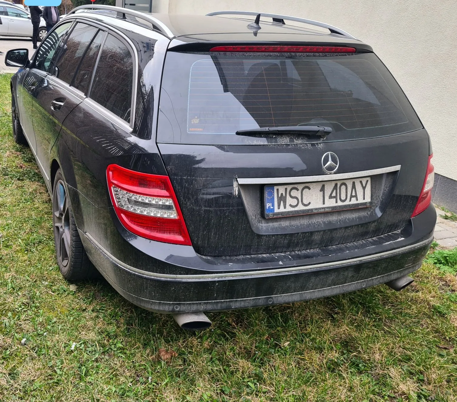Czarny samochód osobowy typu kombi marki Mercedes-Benz zaparkowany na trawie. Widoczny tył pojazdu z logo Mercedes-Benz, czerwonymi światłami tylnymi i tablicą rejestracyjną PL WSC 140AY. Samochód jest zabrudzony, zwłaszcza dolna część nadwozia. W tle budynek z jasną ścianą, fragment innej zaparkowanej osoby oraz trawa z zielonymi i suchymi plamami.