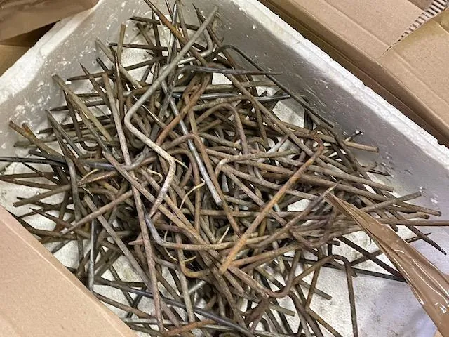 Biały pojemnik z białym podłożem wypełniony zardzewiałymi, zakrzywionymi metalowymi prętami o różnych kształtach i długościach. Pojemnik umieszczony jest wewnątrz kartonowego pudełka. W tle widoczne są fragmenty brązowych kartonów.