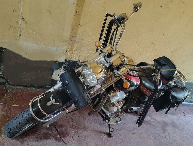 Zdjęcie motocykla typu cruiser w kolorze czarnym i czerwonym, z dużą ilością chromowanych elementów. Motocykl posiada wysoką kierownicę, skórzane torby boczne z frędzlami oraz dodatkowe akcesoria z czarnej skóry przymocowane do przedniego widelca. Pojazd stoi na betonowej posadzce na tle żółtej ściany.