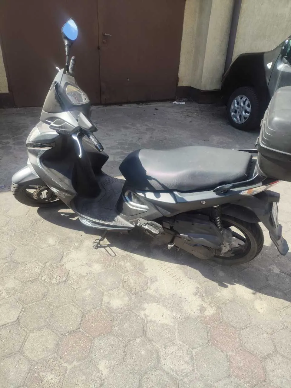 Srebrny skuter zaparkowany na chodniku z sześciokątnymi płytkami. Skuter ma czarne siedzenie, lusterko wsteczne i widoczny napis 'ATMCO' na podłodze. W tle brązowe drzwi garażowe, fragment czarnego samochodu z widocznym kołem oraz budynek z jasną ścianą. Podłoga jest szara z czerwonymi plamami, a na niej leżą drobne śmieci.