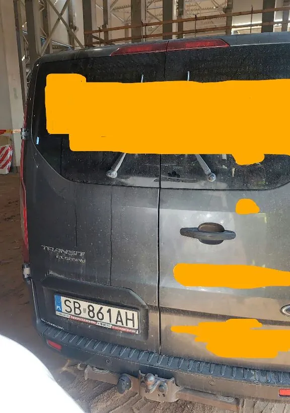 Tylny widok ciemnoszarego samochodu marki Ford Transit Custom. Na klapie bagażnika widoczna jest tablica rejestracyjna o numerze SB 861AH. Część tylnej szyby oraz fragmenty nadwozia są zakryte dużymi, żółtymi plamami. Pojazd znajduje się wewnątrz hali przemysłowej lub magazynu.