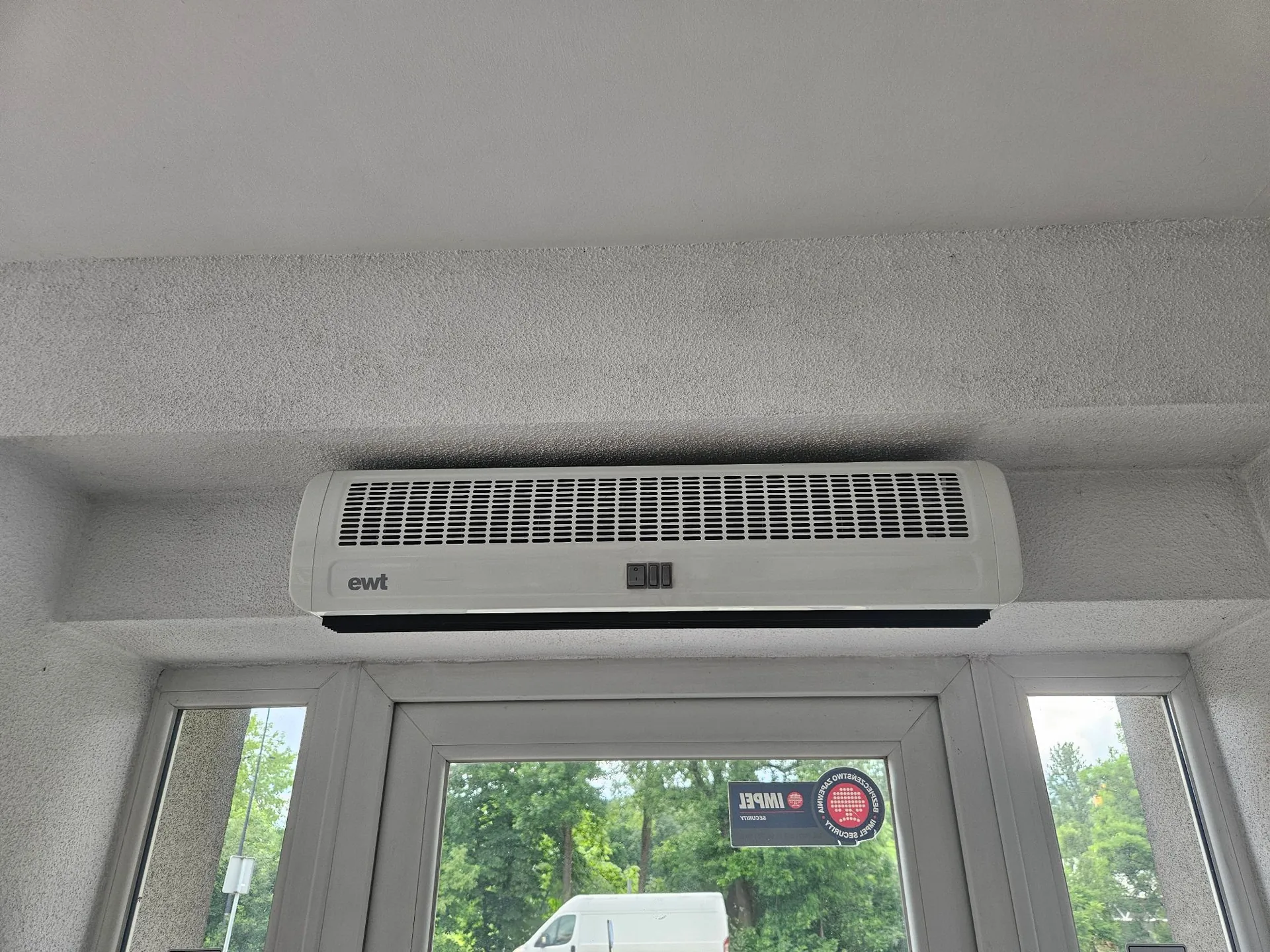 Biała jednostka wewnętrzna klimatyzatora zamontowana na ścianie nad drzwiami. Na obudowie urządzenia widnieje napis 'ewt'. Poniżej znajduje się przeszklone drzwi, na których naklejona jest okrągła naklejka z napisem 'IWBET' oraz dodatkowymi informacjami.