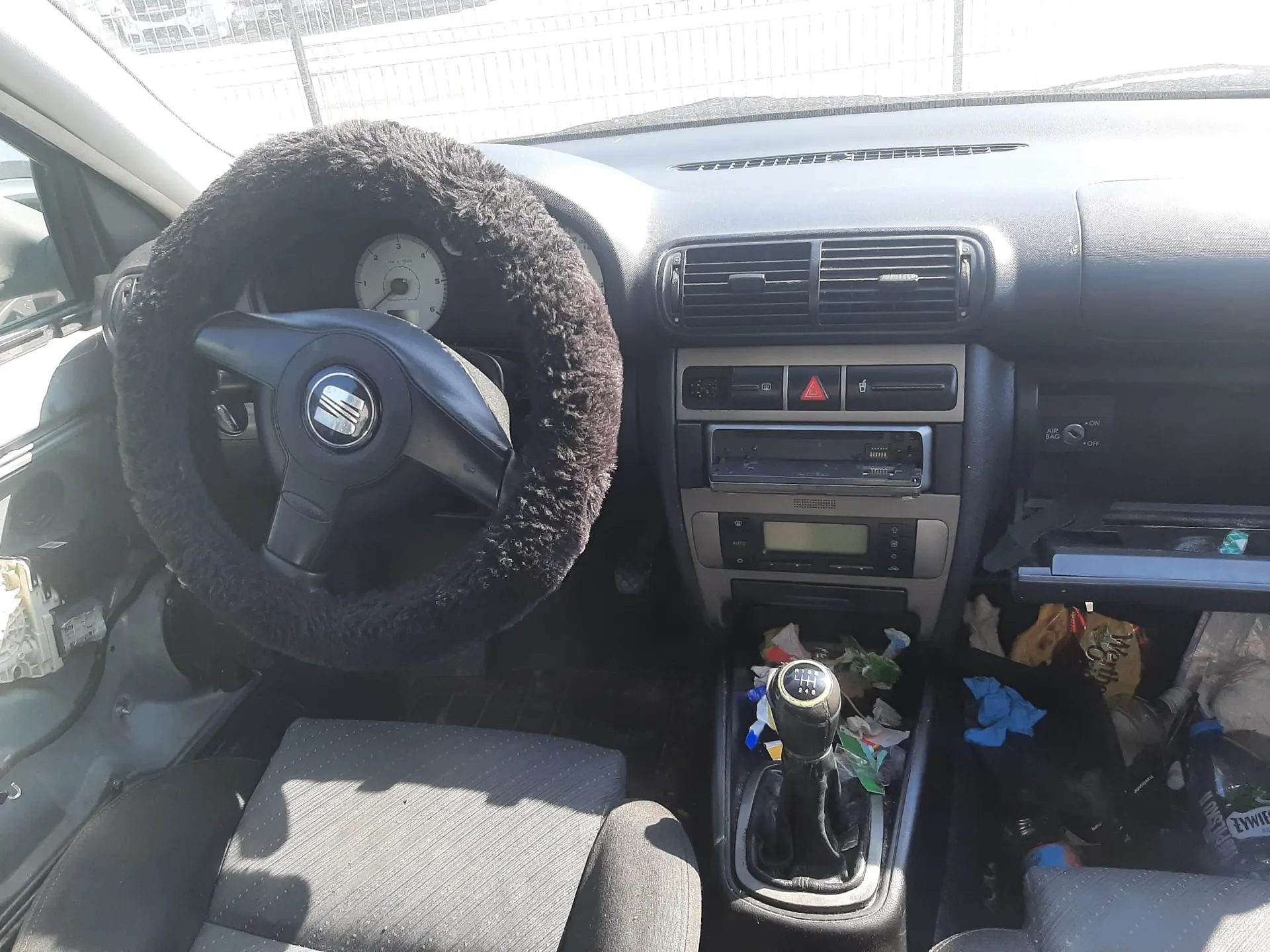 Wnętrze samochodu widziane z perspektywy kierowcy. Kierownica z logo marki SEAT jest owinięta grubym, czarnym, puszystym pokrowcem. Deska rozdzielcza zawiera kratki nawiewów, radio oraz panel sterowania klimatyzacją. W konsoli środkowej znajduje się lewarek zmiany biegów. Podgląd pasażera i podłoga samochodu są zaśmiecone różnymi przedmiotami, w tym plastikowymi opakowaniami i niebieską szmatką.