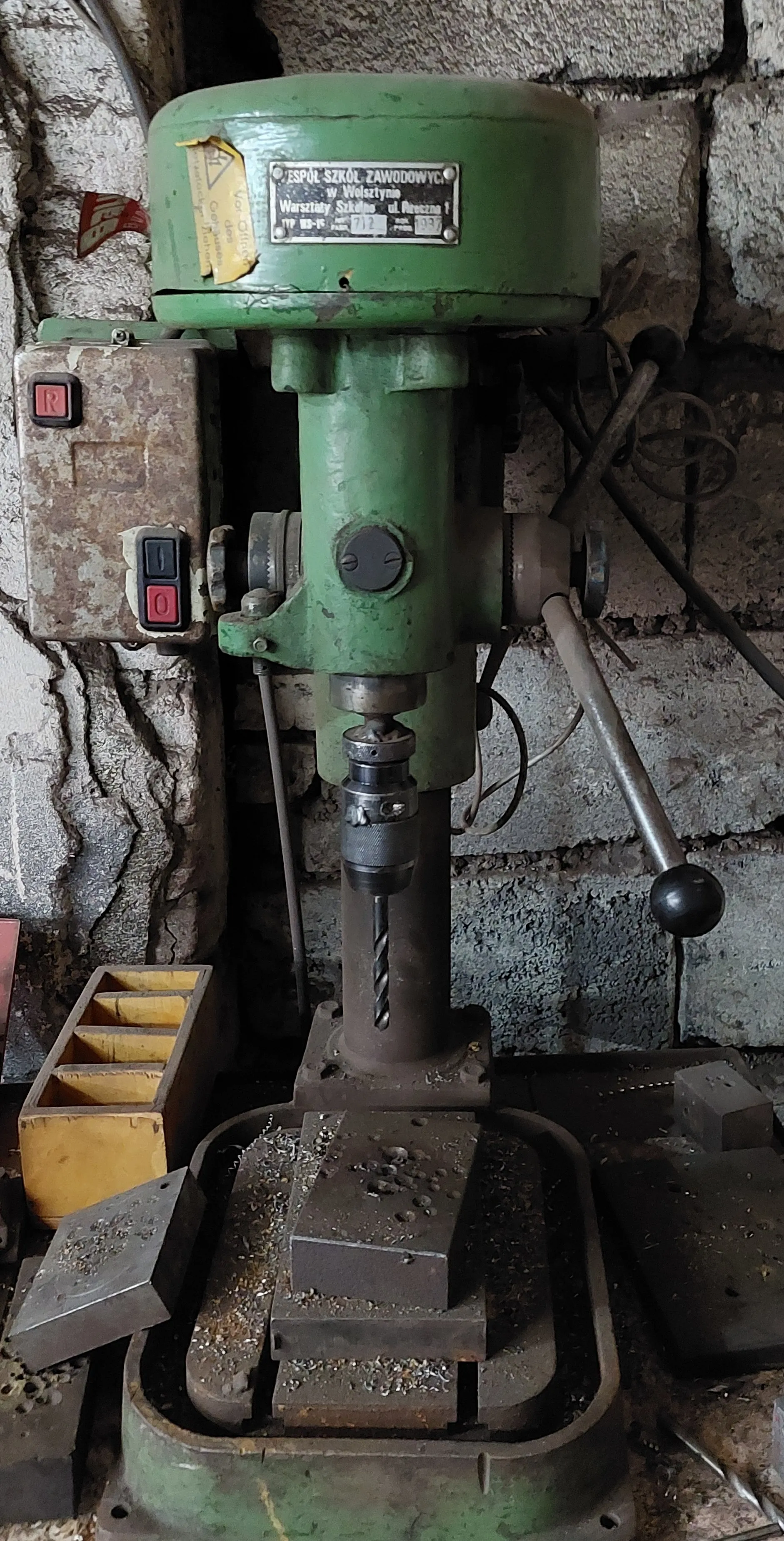 Zielony przemysłowy wiertarka stojąca na metalowej podstawie z metalowymi wiórami. Na maszynie tabliczka z napisem 'ZESPÓŁ SZKÓŁ ZAWODOWYCH w Wolsztynie Warsztaty Szkolne ul. Racławicka 1' oraz numerami '712' i '1957'. Po lewej stronie panel sterowania z czerwonym przyciskiem 'R' i przyciskami 'I' oraz 'O'. Tło stanowi betonowa ściana z pęknięciami. Obok wiertarki żółte pudełko z przegródkami i metalowe elementy.