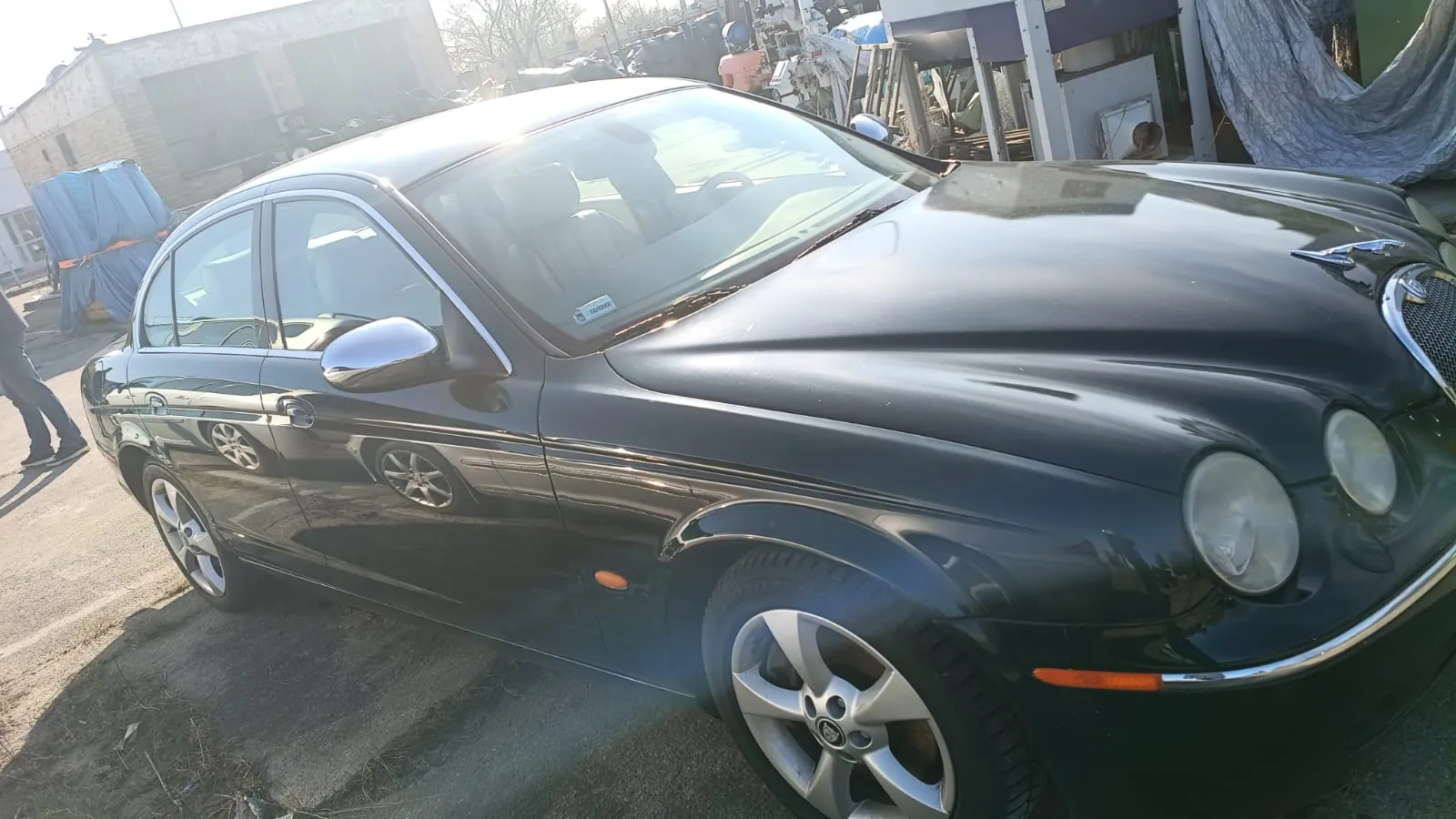 Czarny sedan marki Jaguar zaparkowany na zewnątrz. Samochód ma błyszczącą karoserię, srebrne felgi i widoczne reflektory. W tle budynki, przedmioty przykryte niebieskimi i szarymi tarpami oraz osoba idąca po lewej stronie. Słońce oświetla przednią część pojazdu, powodując połysk na masce.