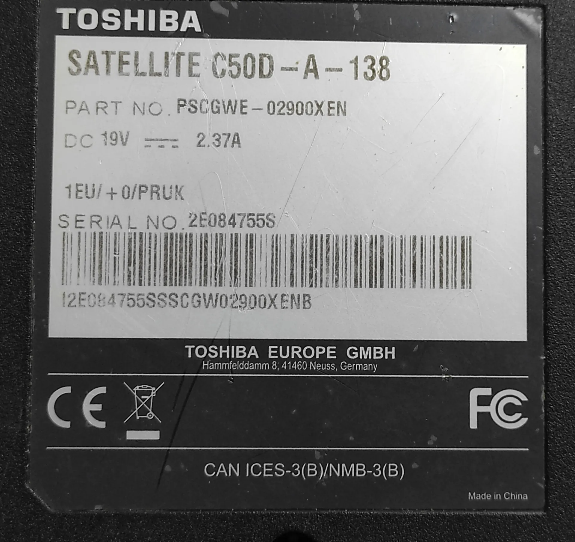 Zdjęcie tabliczki znamionowej laptopa marki Toshiba. Na białym tle widnieją napisy: 'TOSHIBA SATELLITE C50D - A - 138', 'PART NO. PSCGWE - 02900XEN', 'DC 19V 2.37A', '1EU/ + 0/PRUK', 'SERIAL NO. 2E084755S'. Poniżej znajduje się kod kreskowy z numerem '12E084755SSSCGW02900XENB'. Na dole tabliczki widnieje napis 'TOSHIBA EUROPE GMBH Hammfelddamm 8, 41460 Neuss, Germany', znaki certyfikacji CE i FC, napis 'CAN ICES-3(B)/NMB-3(B)' oraz 'Made in China'.