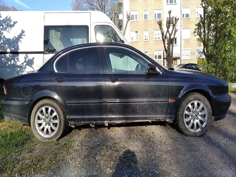 Czarny samochód osobowy typu sedan zaparkowany na żwirowym podłożu. Po lewej stronie widoczny biały furgon, w tle wielopiętrowy blok mieszkalny z oknami oraz drzewa. Na pierwszym planie trawa i cień od drzewa. Niebo jest jasne, słoneczna pogoda.