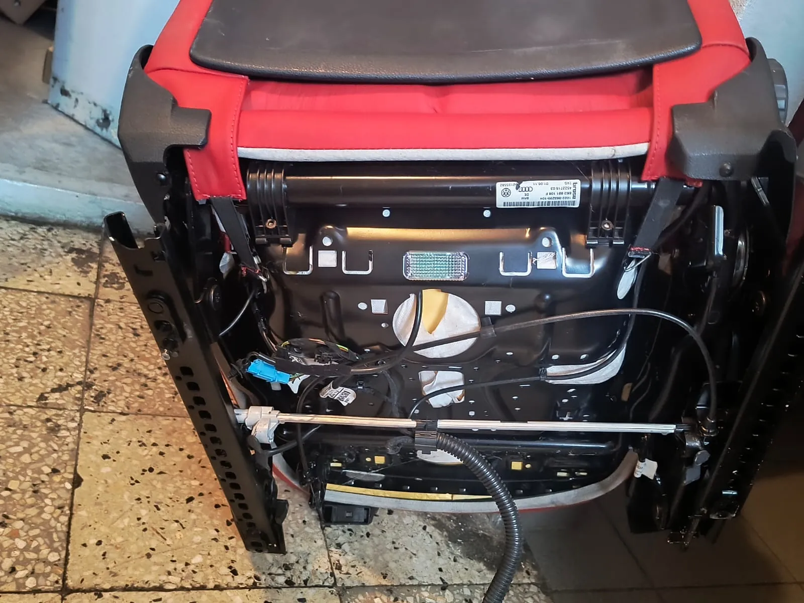 Zdjęcie przedstawiające tył demontowanego fotela samochodowego z czerwonym obiciem i czarnym zagłówkiem. Widoczna jest metalowa konstrukcja ramy, wiązki przewodów elektrycznych oraz biała etykieta z logo Audi i Volkswagen oraz kodami kreskowymi. Fotel spoczywa na brudnej, kafelkowej podłodze.