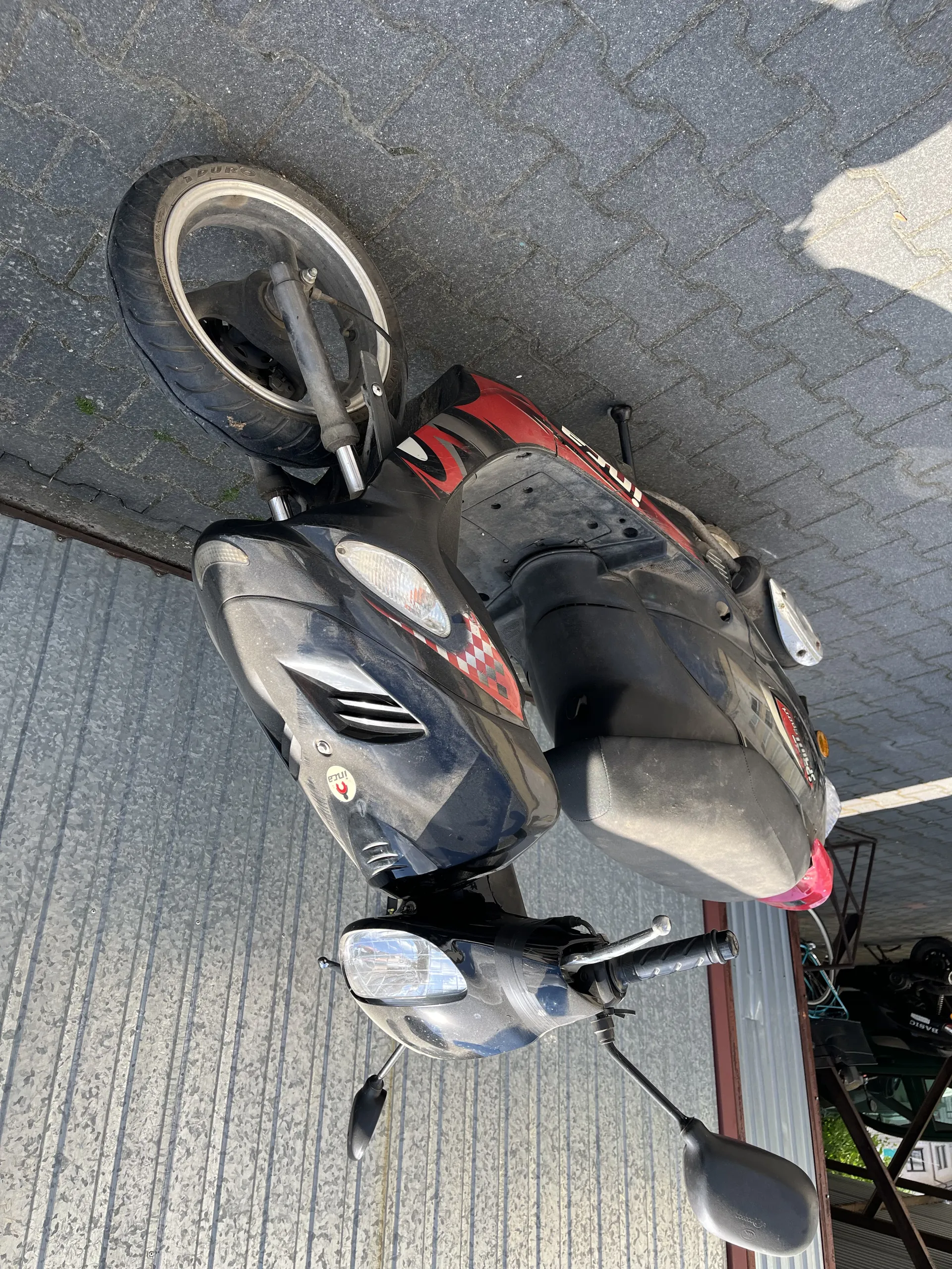 Czarny skuter z czerwonymi i białymi szachownicowymi wzorami po bokach, zaparkowany na brukowanej powierzchni. W tle metalowa ściana z pionowymi liniami. Skuter posiada dwa lusterka, reflektor i siedzenie. Na przedniej części widoczny mały okrągły naklejony znaczek.