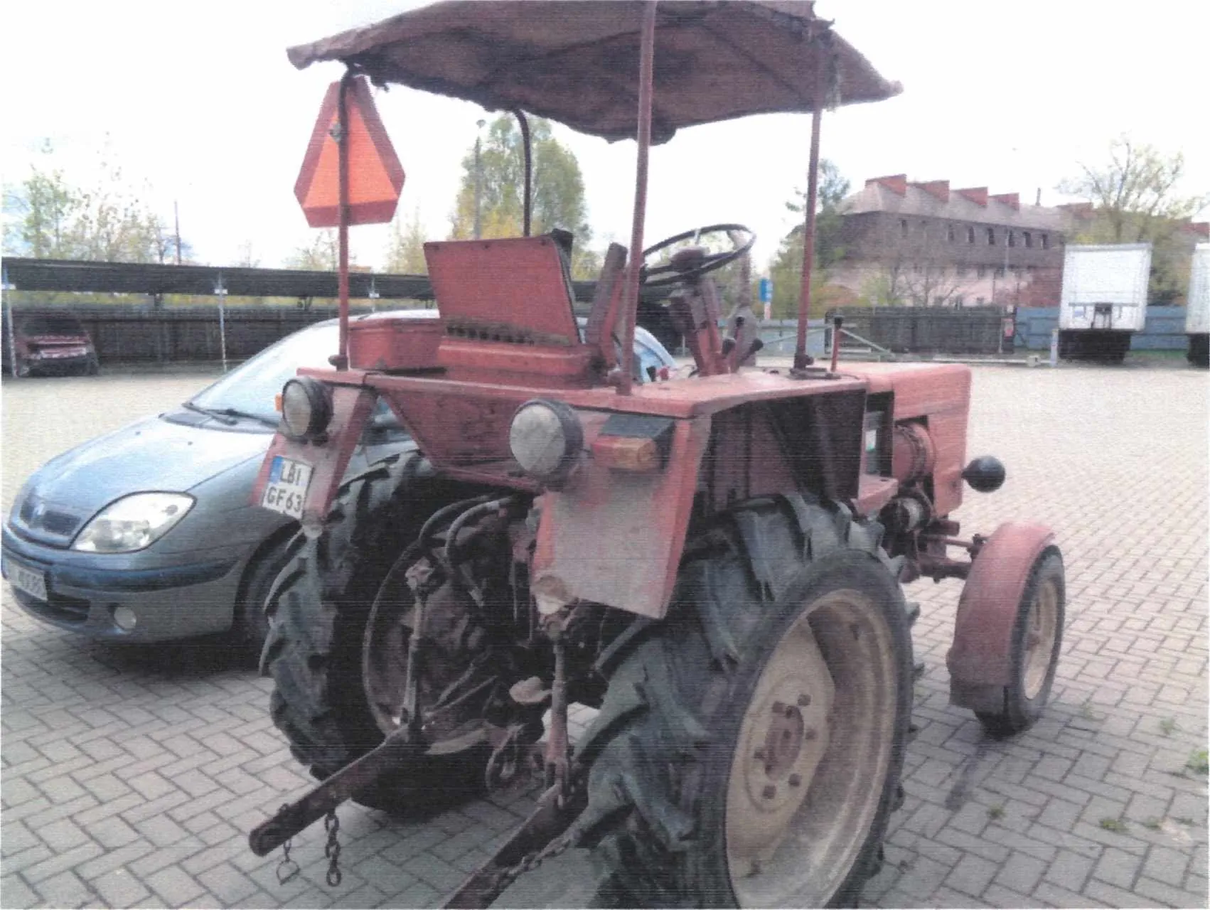 Czerwony traktor z daszkiem, widok od tyłu. Na traktorze widoczna tablica rejestracyjna LBI GF63 oraz pomarańczowy trójkąt ostrzegawczy. W tle szary samochód osobowy, budynki, drzewa i białe ciężarówki na brukowanym placu. Traktor ma duże opony, kierownicę i metalowe elementy. Niebo jest pochmurne.