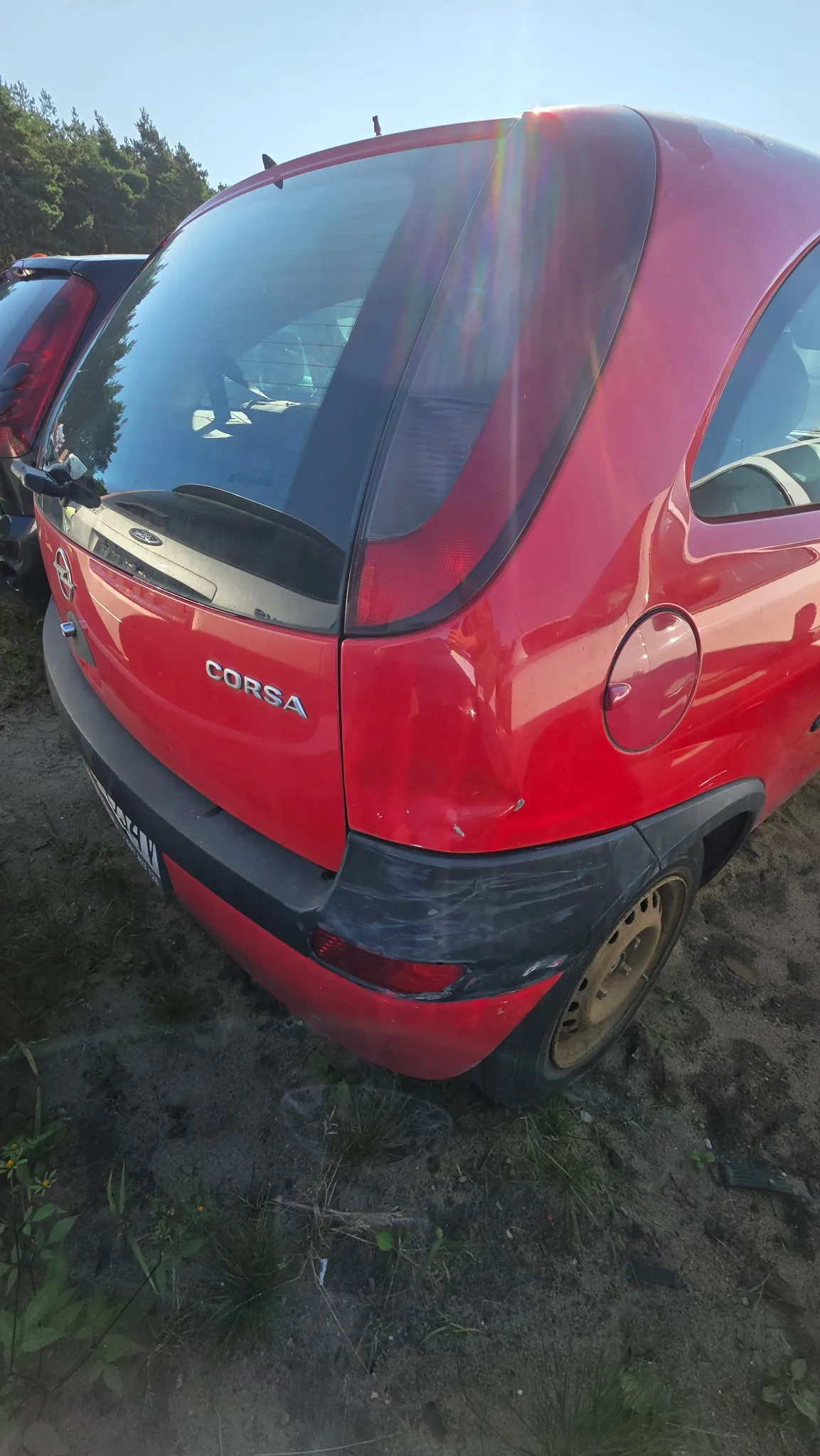 Tylna część czerwonego samochodu marki Opel Corsa. Pojazd posiada widoczne uszkodzenia w postaci wgniecenia na tylnym błotniku po prawej stronie oraz zarysowania na czarnym plastikowym zderzaku. Na klapie bagażnika widnieje srebrny napis CORSA oraz logo marki Opel. Samochód stoi na piaszczystym podłożu.