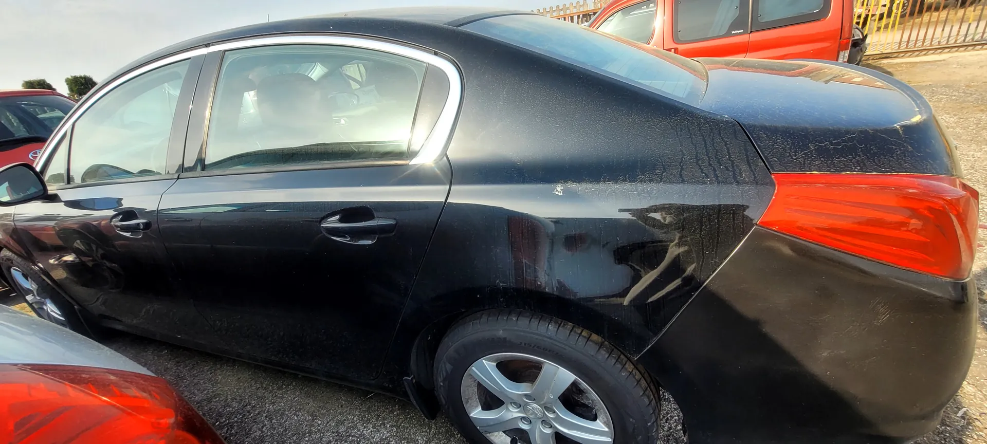 Czarny samochód osobowy typu sedan widziany z boku i tyłu, z matowym lakierem pokrytym drobnymi zadrapaniami. Widoczne są przyciemniane szyby, srebrna listwa nad drzwiami, czerwone światła tylne oraz felgi ze srebrnymi dyskami. W tle częściowo widoczny czerwony pojazd, metalowe ogrodzenie i asfaltowy parking. Niebo jest jasne, bez chmur.