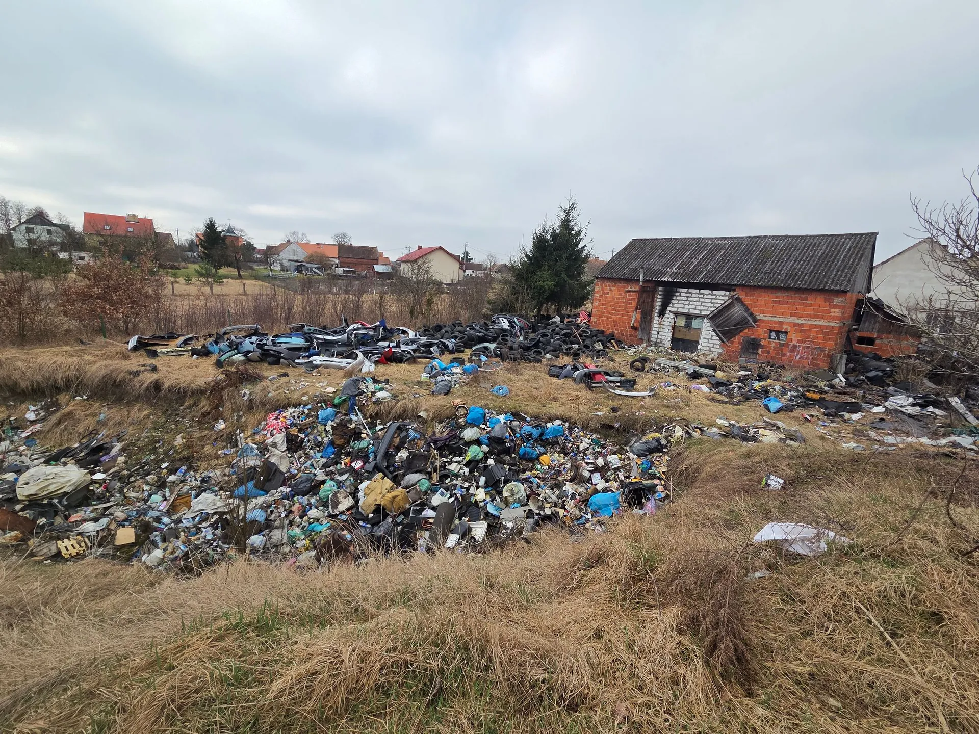 Zdjęcie przedstawiające dzikie wysypisko śmieci na trawiastym terenie. Na pierwszym planie i w centrum widoczne są duże sterty odpadów, w tym plastikowe butelki, fragmenty opon, metalowe elementy i inne odpady komunalne. W tle po prawej stronie znajduje się niewielki, częściowo zniszczony budynek z czerwonej cegły z dziurawym dachem. W oddali widoczne są domy jednorodzinne oraz drzewa pod zachmurzonym niebem.