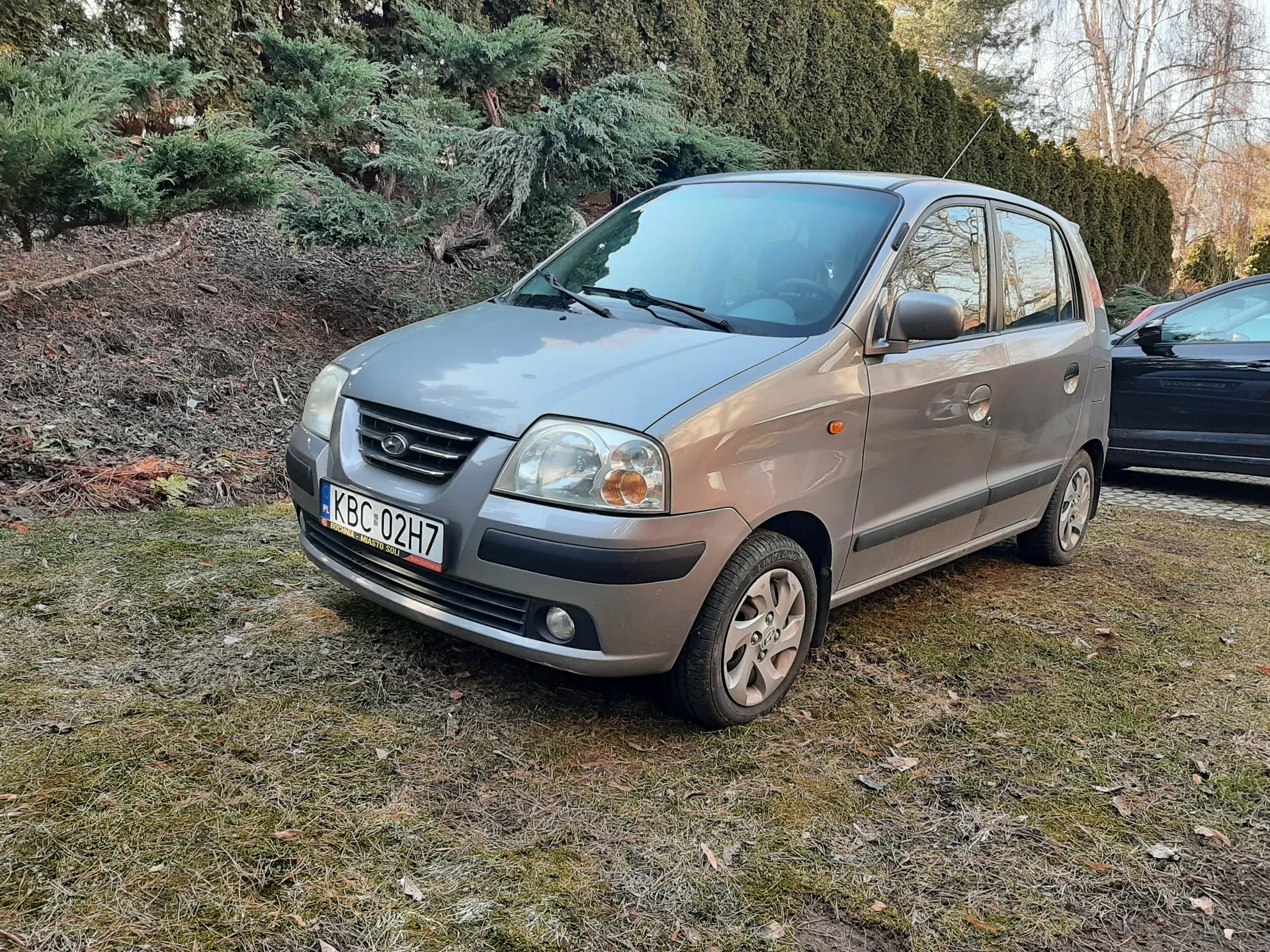 Srebrny samochód osobowy marki Daewoo Matiz zaparkowany na trawie. Widoczna tablica rejestracyjna KBC 02H7 z napisem "PL" oraz dodatkowym napisem "MIASTO SŁUPSK" na dole. W tle zielone krzewy i drzewa, po prawej stronie częściowo widoczny czarny samochód. Trawa na podwórku jest wyschnięta z miejscami zielonej trawy.