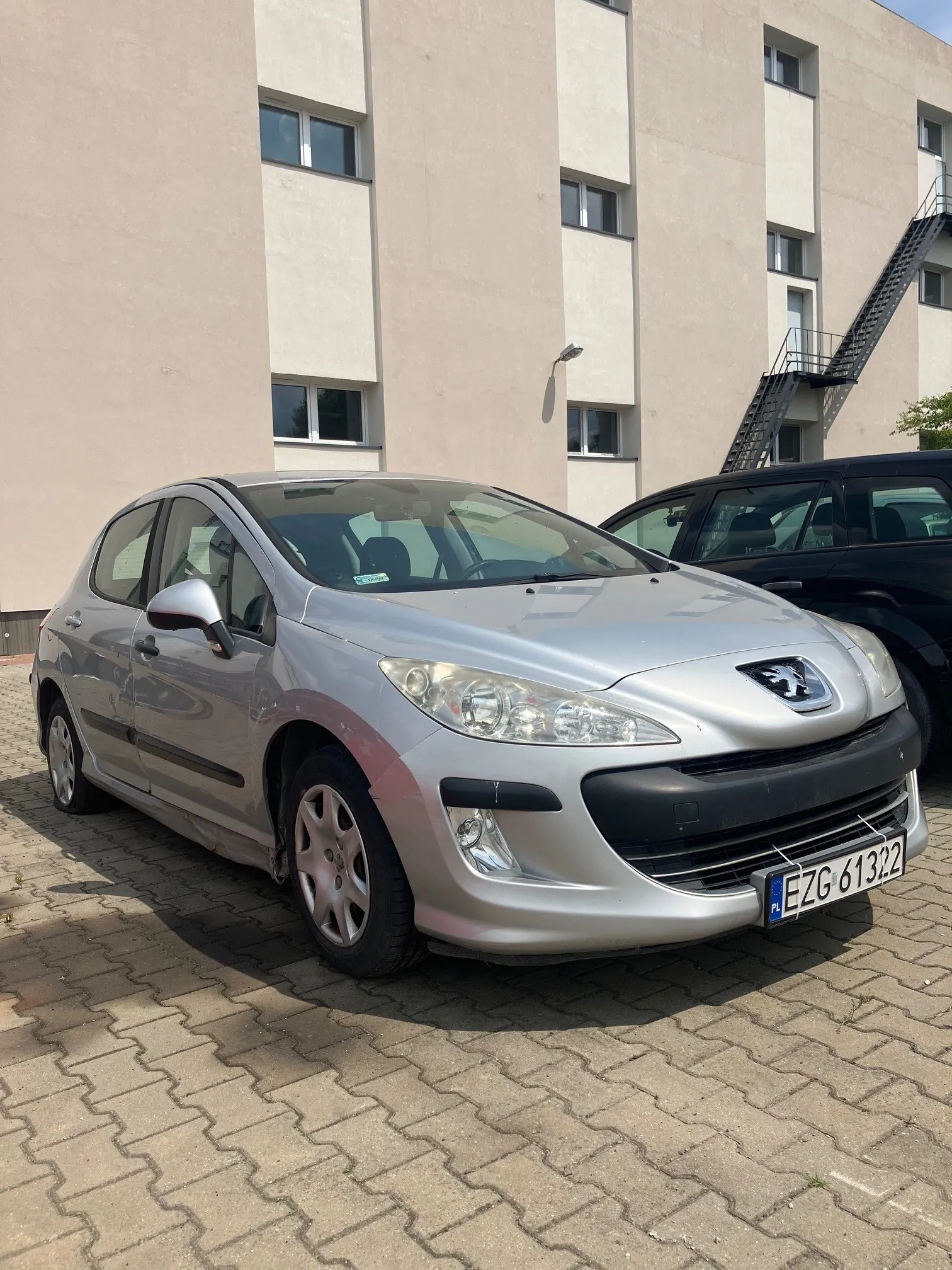 Srebrny samochód osobowy marki Peugeot zaparkowany na kostce brukowej przed jasnym, wielopiętrowym budynkiem. Pojazd ma polską tablicę rejestracyjną o numerze EZG 61322. W tle widoczny jest fragment innego, ciemnego samochodu oraz zewnętrzne schody przeciwpożarowe przy ścianie budynku.