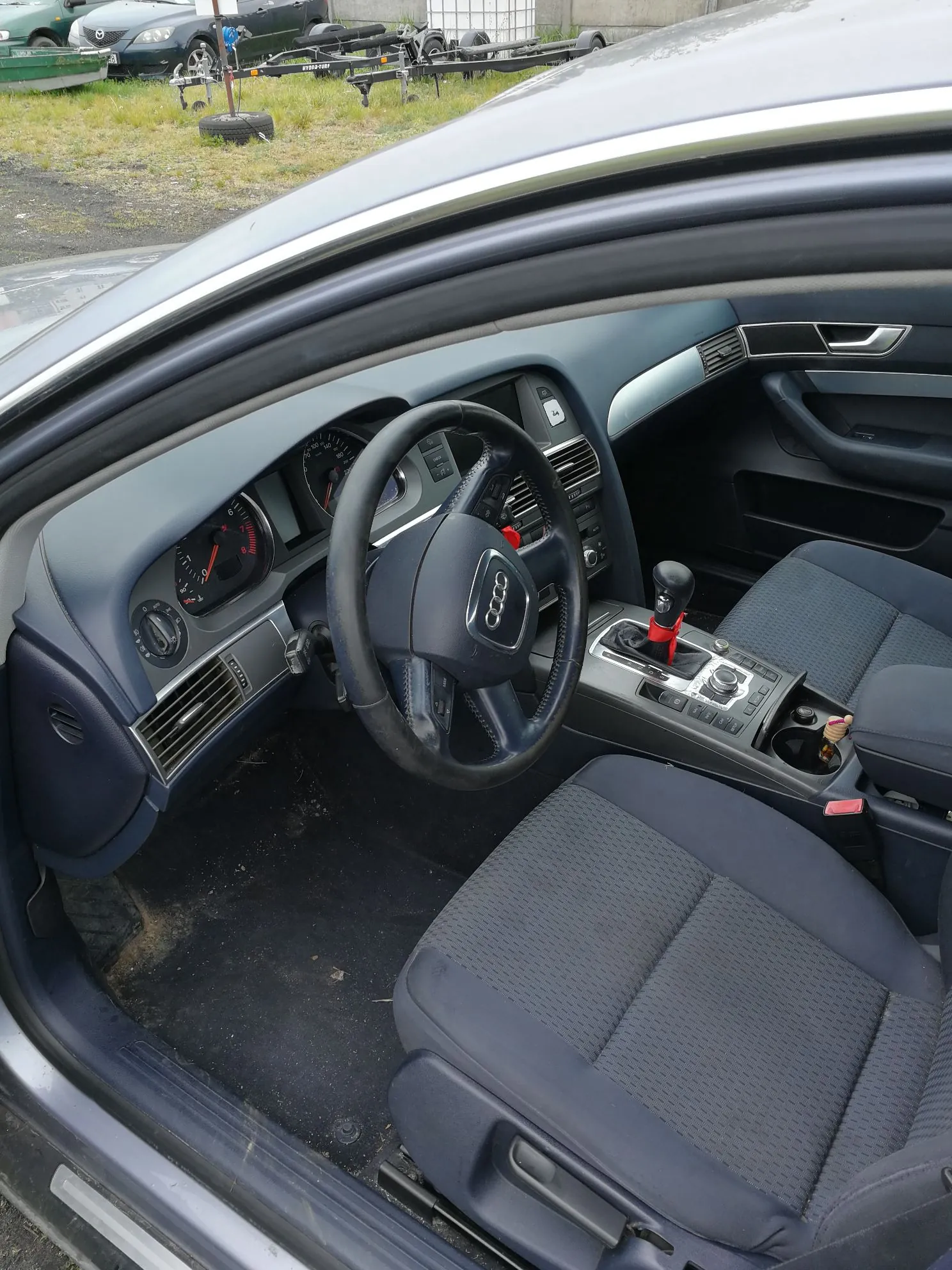 Wnętrze samochodu widziane z otwartych drzwi kierowcy. Widoczny jest ciemnoszary fotel z materiałową tapicerką, czarna kierownica z logo Audi, deska rozdzielcza z zegarami, konsola środkowa z lewarkiem zmiany biegów oraz fragmenty drzwi i podsufitki. W tle, za szybą, widoczne są inne samochody i przyczepy na placu.