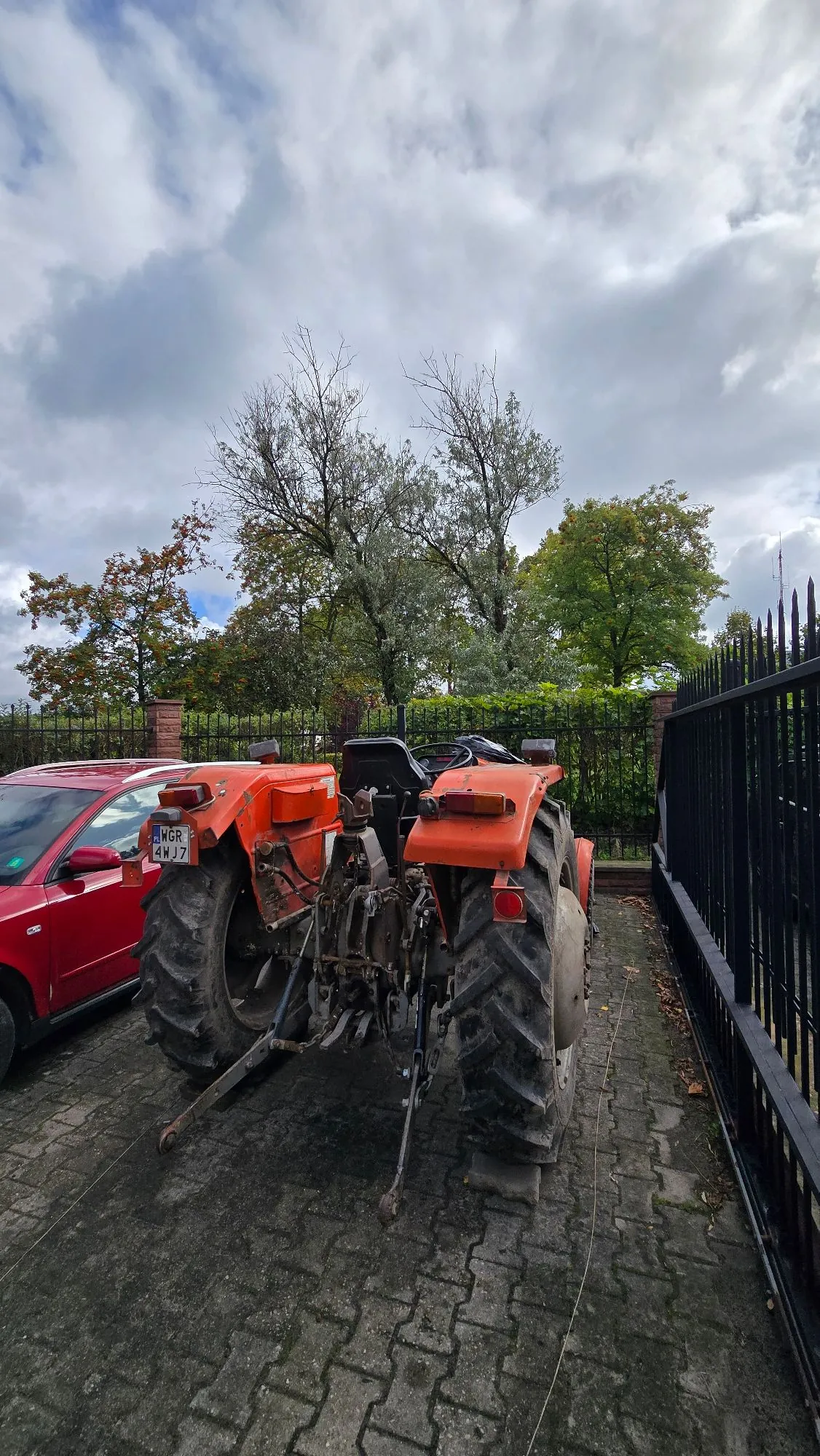 Widok z tyłu na pomarańczowy traktor zaparkowany na kostce brukowej obok czerwonego samochodu osobowego. Traktor posiada duże, błotniste koła z wyraźnym bieżnikiem oraz układ hydrauliczny z tyłu. Po prawej stronie znajduje się czarny, metalowy płot, a w tle zielone drzewa i zachmurzone niebo. Na czerwonym samochodzie widoczna jest tablica rejestracyjna o numerze WGR 4WJ7.