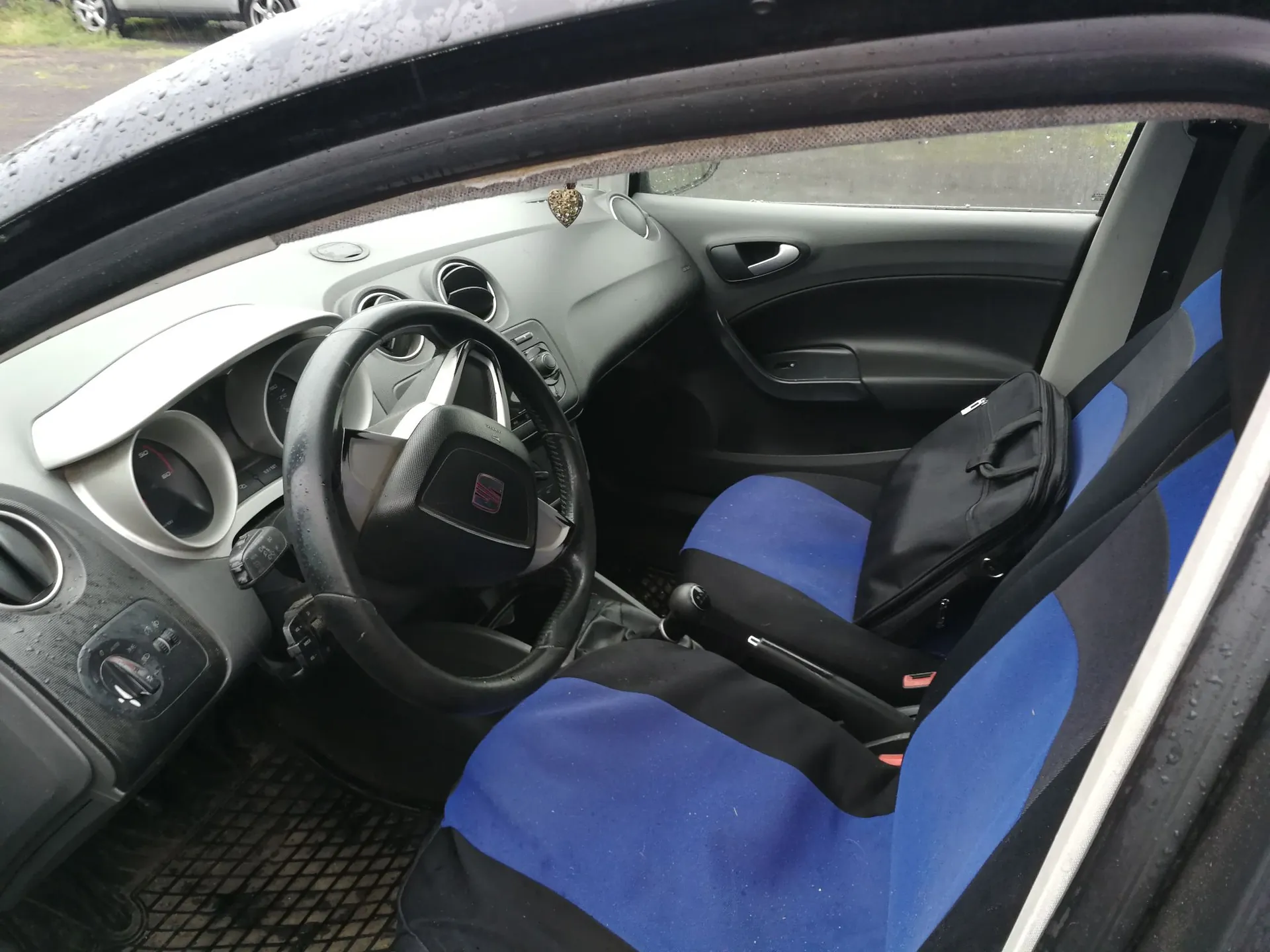 Wnętrze samochodu marki Seat widziane z perspektywy otwartych drzwi kierowcy. Widoczna jest czarna kierownica z logo Seat, deska rozdzielcza, wskaźniki oraz fotel kierowcy z niebiesko-czarną nakładką materiałową. Na siedzeniu leży czarna torba. Zdjęcie wykonano w deszczowy dzień, co widać po kroplach wody na ramie drzwi.