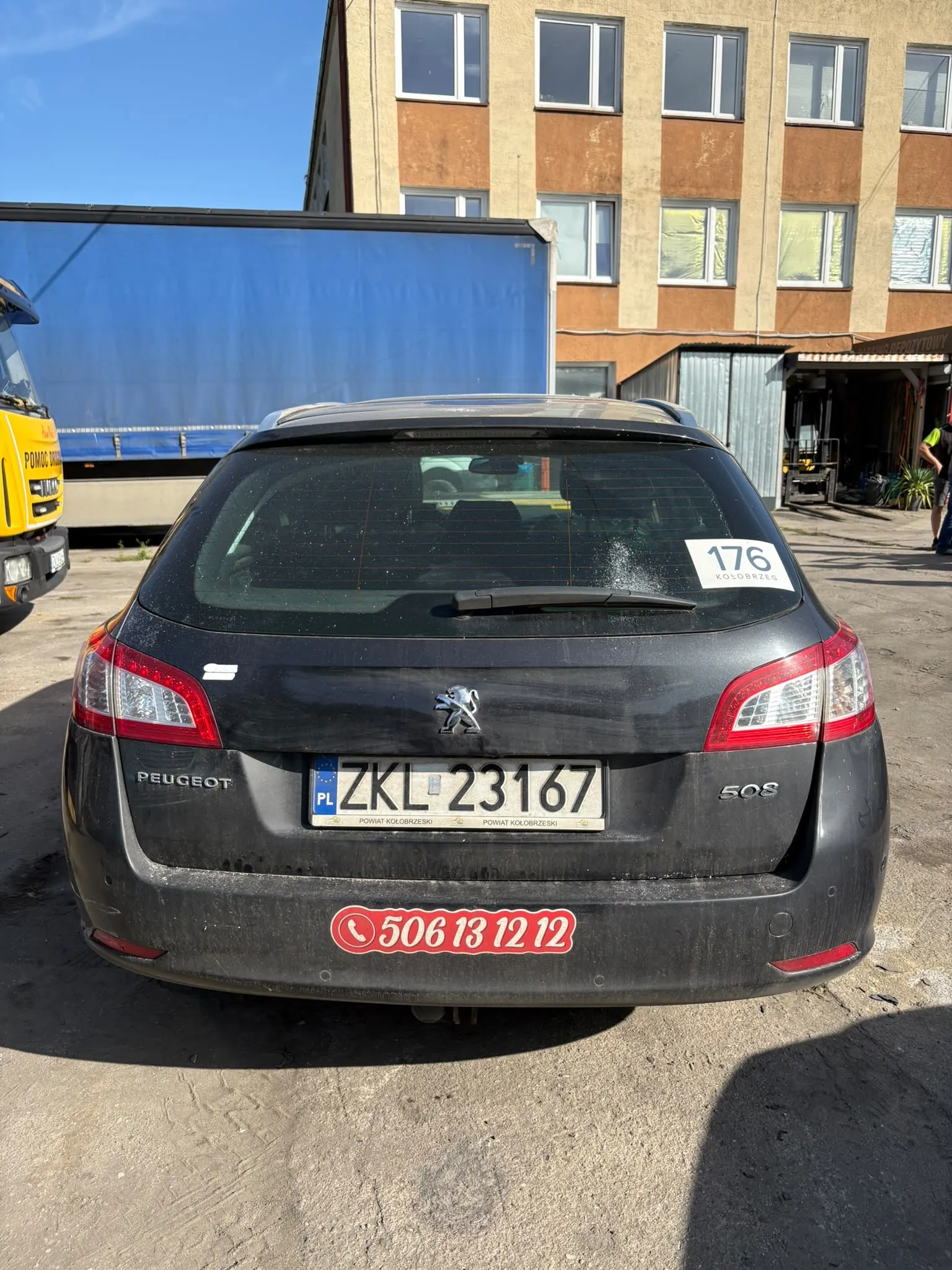 Tylny widok czarnego samochodu marki Peugeot 508 zaparkowanego na betonowym placu. Samochód ma polską tablicę rejestracyjną ZKL 23167 oraz czerwoną naklejkę z numerem telefonu 506 13 12 12 na zderzaku. W prawym górnym rogu tylnej szyby znajduje się biała naklejka z numerem 176. W tle widoczna jest niebieska plandeka ciężarówki oraz wielopiętrowy budynek o żółto-brązowej elewacji z wieloma oknami.
