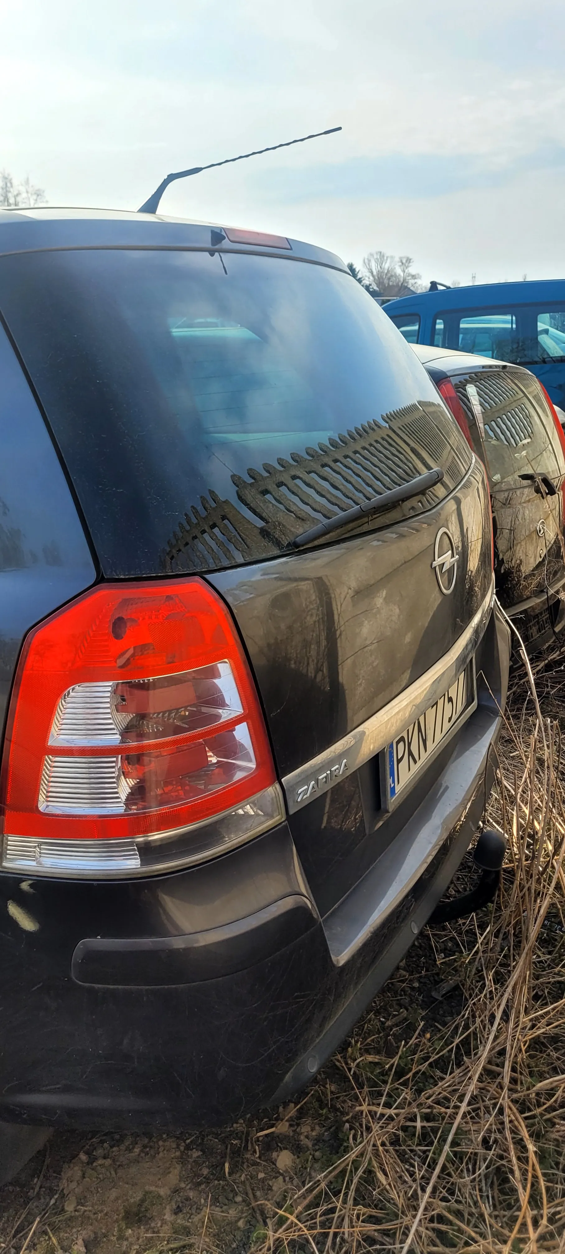 Zdjęcie przedstawia tył czarnego samochodu Opel Zafira z widocznym napisem "ZAFIRA" po lewej stronie bagażnika. Na samochodzie znajduje się czerwone światło tylnego reflektora, czarna osłona szyby z wycieraczką oraz tablica rejestracyjna o numerze PKN 775L z niebieskim pasem po lewej stronie. W tle widoczne są inne pojazdy, suche trawy na ziemi oraz fragment nieba z chmurami. W odbiciu na szybie bagażnika widać drewniany płot.