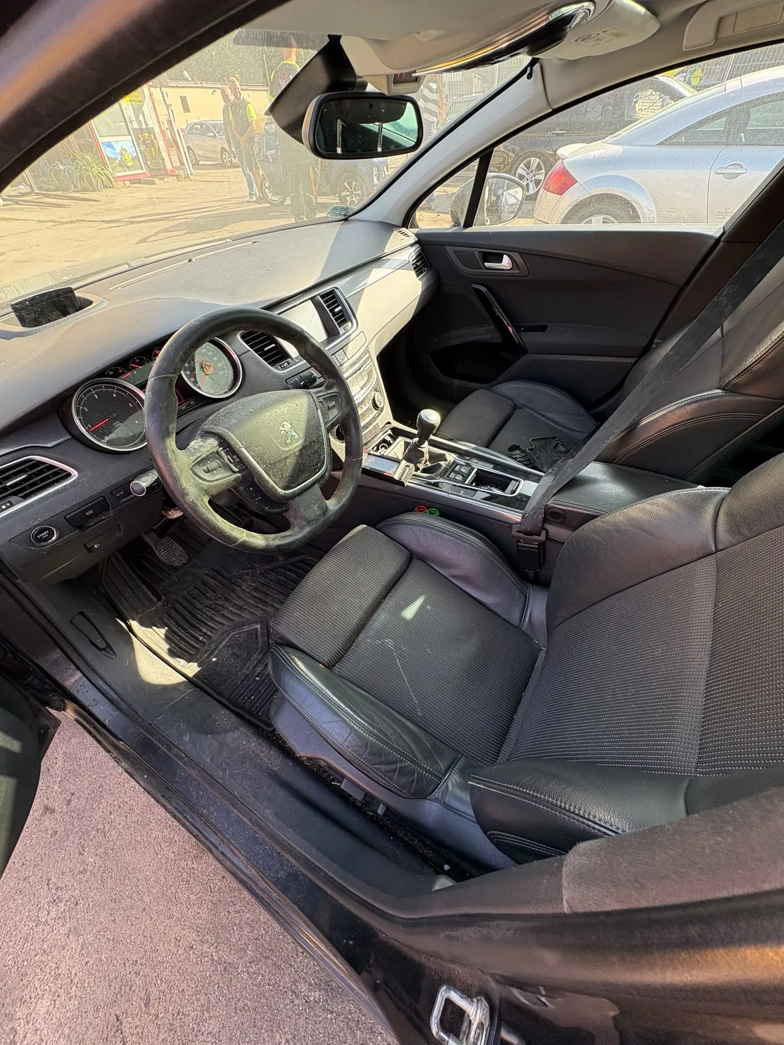 Wnętrze samochodu marki Peugeot widziane z perspektywy otwartych przednich drzwi kierowcy. Widoczna jest czarna skórzana tapicerka foteli, kierownica z logo marki, deska rozdzielcza z zegarami analogowymi oraz konsola środkowa z lewarkiem zmiany biegów. Przez przednią szybę i okno widać fragmenty innych samochodów zaparkowanych na zewnątrz w słoneczny dzień.