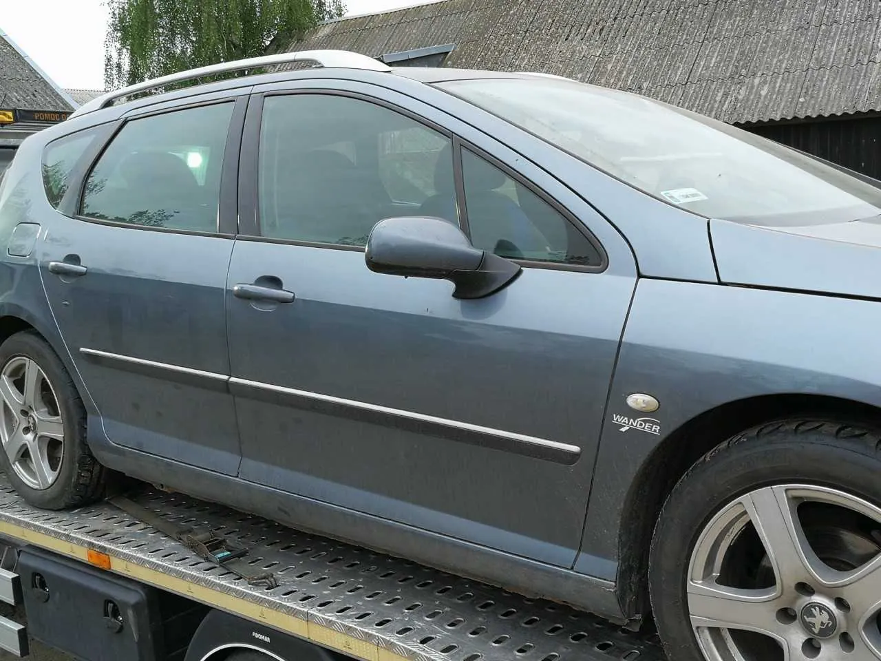 Szary samochód osobowy typu kombi z napisem "WANDER" na przednim błotniku, umieszczony na płycie załadowczej holownika. Widoczne są srebrne felgi, dachowy bagażnik i lusterko boczne. W tle budynek z blachą falistą, drzewa oraz tablica z częściowo widocznym napisem "POMOC". Samochód i elementy tła są ustawione poziomo, bez oznak nieprawidłowej orientacji.
