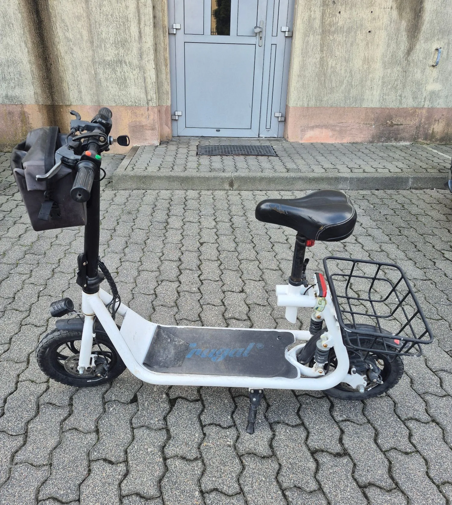 Biała hulajnoga elektryczna marki Regal stojąca na kostce brukowej przed szarymi drzwiami budynku. Hulajnoga posiada czarną torbę zamontowaną na kierownicy, czarne siedzisko oraz czarny koszyk bagażowy z tyłu.
