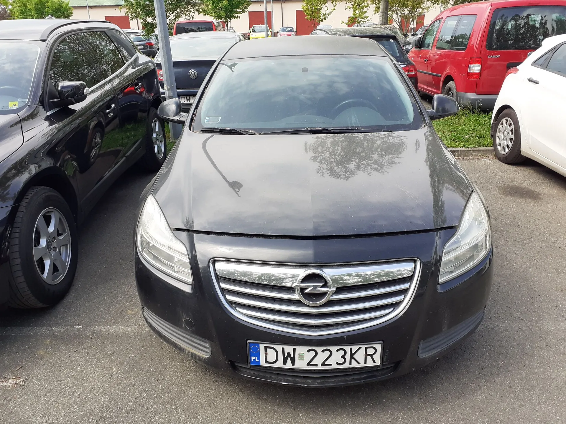 Czarny samochód marki Opel zaparkowany na parkingu asfaltowym. Widoczna jest przednia część pojazdu z tablicą rejestracyjną o numerze DW 223KR. Po lewej stronie stoi inny czarny samochód, a w tle czerwony furgon oraz biały samochód osobowy.