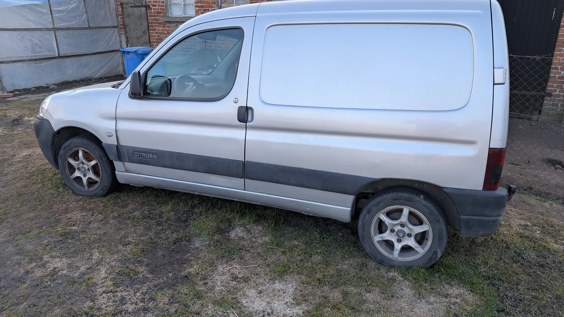 Srebrny samochód dostawczy Citroën zaparkowany na trawie z widocznym napisem CITROËN na boku. W tle ceglany budynek z oknem, niebieski pojemnik na śmieci, konstrukcja zasłonięta folią oraz siatka ogrodzeniowa. Samochód ma czarne pasy na bokach i srebrne felgi.