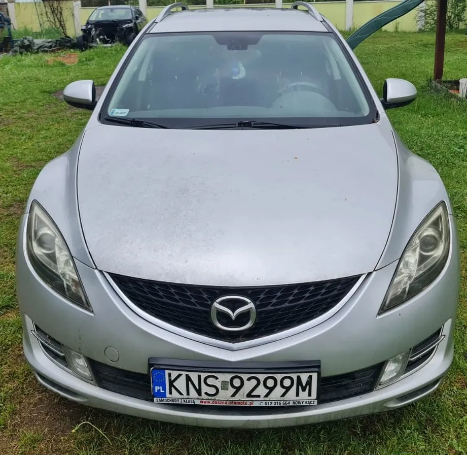 Przedni widok srebrnego samochodu marki Mazda z tablicą rejestracyjną KNS 9299M. Na tablicy widoczny jest napis 'PL' oraz dodatkowy tekst: 'SAMOCHODY Z KLASY www.doszna.automoto.pl 512 316 664 NOWY SĄCZ'. W tle zielona trawa, uszkodzony czarny samochód oraz żółty budynek z białymi słupkami. Samochód stoi na trawie, a na przedniej szybie widoczny jest niebieski znaczek.