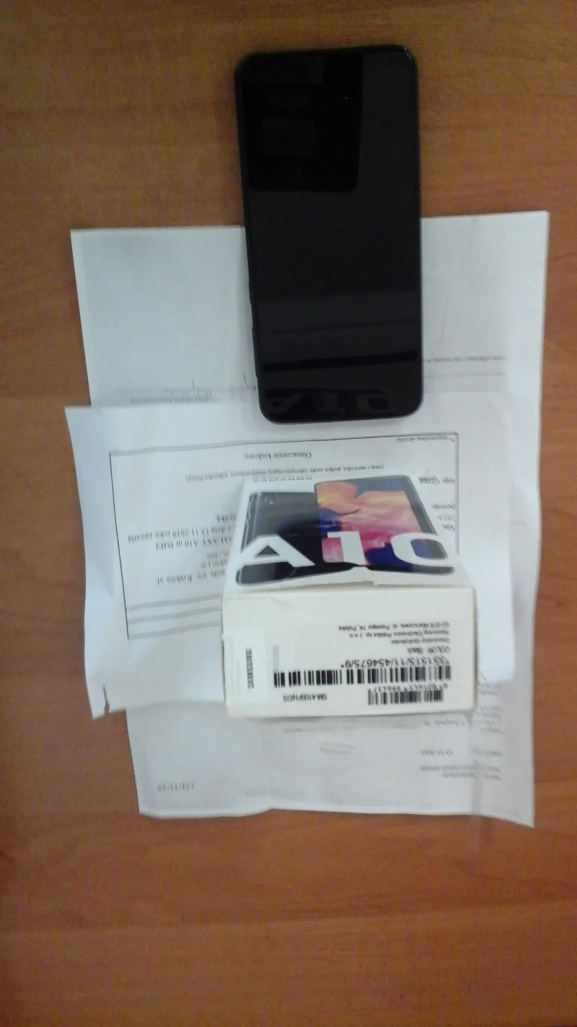 Na drewnianym stole leżą białe dokumenty z nieczytelnym tekstem, pudełko Samsung A10 z kodem kreskowym i czarny smartfon. Na pudełku widoczne są napisy 'SAMSUNG' oraz 'A10'.