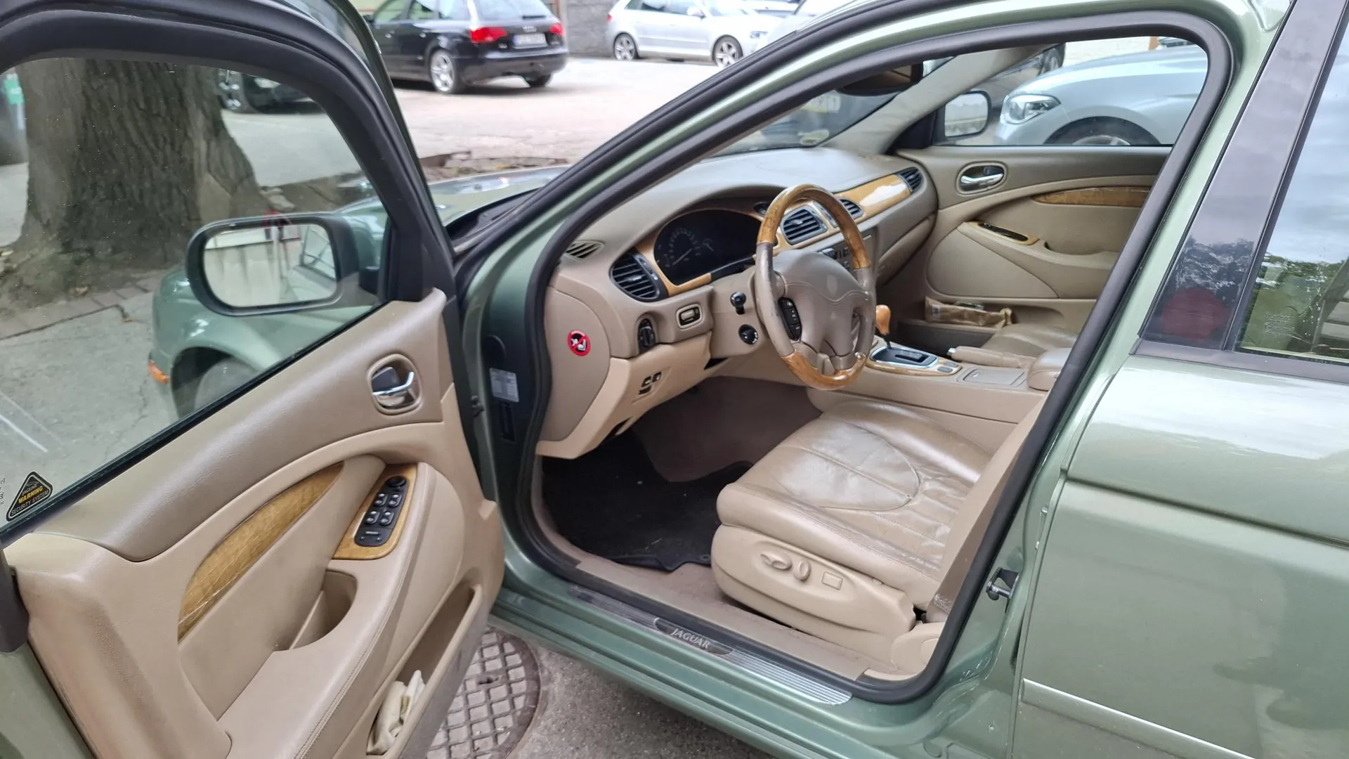 Wnętrze samochodu marki Jaguar w kolorze beżowym, widziane z otwartych drzwi kierowcy. Widoczna jest kierownica z drewnianymi elementami, skórzane fotele, deska rozdzielcza oraz panel boczny drzwi z drewnianą wstawką. Samochód ma lakier w kolorze jasnozielonym. Na progu samochodu widoczny jest napis 'JAGUAR'. W tle widać inne zaparkowane samochody.