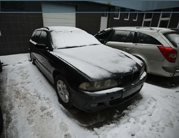 Dwa samochody zaparkowane na śnieżnym podwórku. Czarny samochód osobowy typu kombi z przodu, pokryty warstwą śniegu na dachu i masce. Obok niego srebrny samochód osobowy, również częściowo pokryty śniegiem. Tło stanowi budynek z ciemnymi płytkami i białymi drzwiami garażowymi. Na ziemi widoczne ślady po stopach w śniegu.