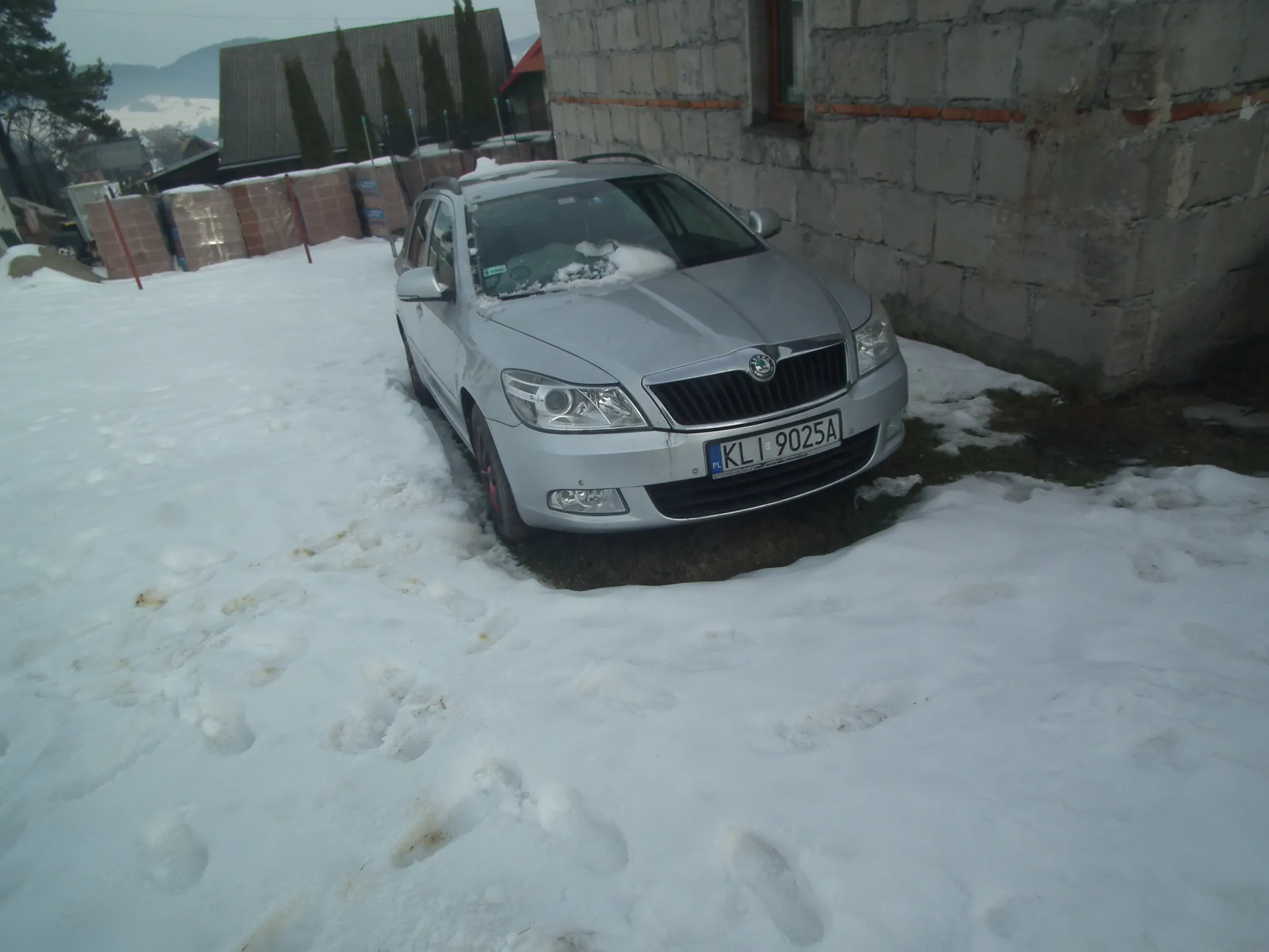 Srebrny samochód marki Škoda z tablicą rejestracyjną KL 9025A zaparkowany na śnieżnej powierzchni z widocznymi śladami stóp. Po prawej stronie kamienna ściana budynku, w tle stosy cegieł, drzewa i góry. Na dachu i przedniej szybie samochodu leży śnieg.