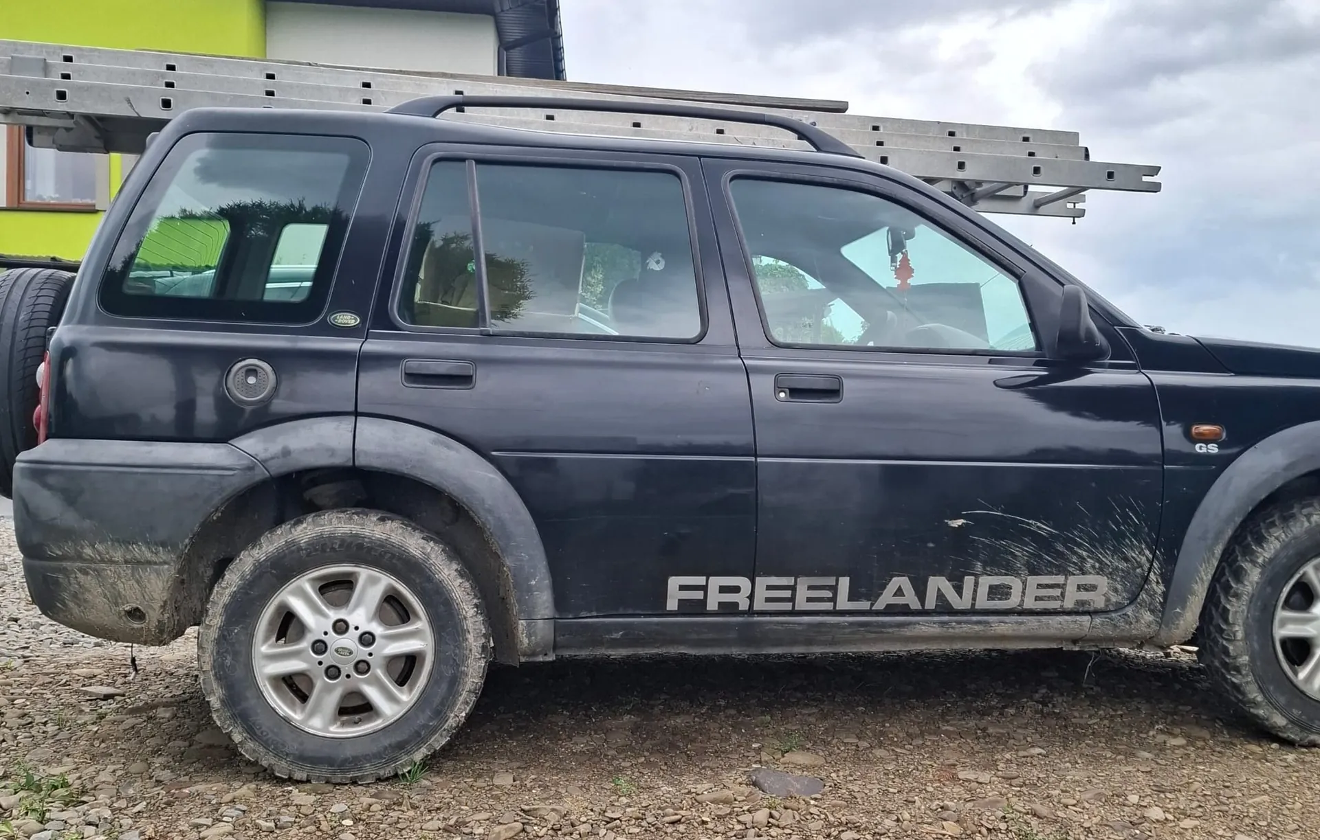 Czarny samochód terenowy marki Land Rover Freelander zaparkowany na żwirowym podłożu. Pojazd jest widoczny z profilu, z boku widnieje duży napis 'FREELANDER'. Na dachu samochodu zamontowany jest metalowy stelaż z drabiną. W tle widać fragment jasnozielonego budynku.