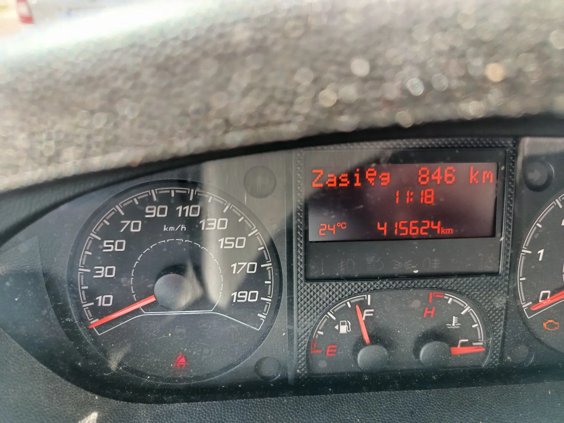 Zbliżenie na deskę rozdzielczą samochodu. Po lewej stronie znajduje się prędkościomierz z zaznaczonymi wartościami od 10 do 190 km/h. W centralnej części widoczny jest czerwony wyświetlacz cyfrowy z napisami: 'Zasięg 846 km', godzina '11:18', temperatura '24°C' oraz przebieg '415624 km'. Poniżej wyświetlacza znajdują się wskaźniki poziomu paliwa oraz temperatury cieczy chłodzącej.