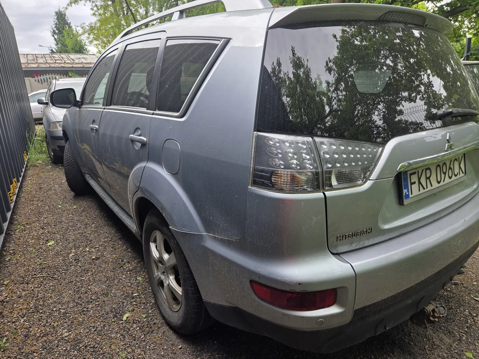Srebrny SUV marki Mitsubishi zaparkowany na żwirowej powierzchni. Widoczny tył pojazdu z tablicą rejestracyjną PL FKR 096CKI, reflektorami i napisem "MITSUBISHI". Po lewej stronie metalowy płot z żółtymi znakami, w tle drugi samochód i drzewa. Niebo jest zachmurzone.