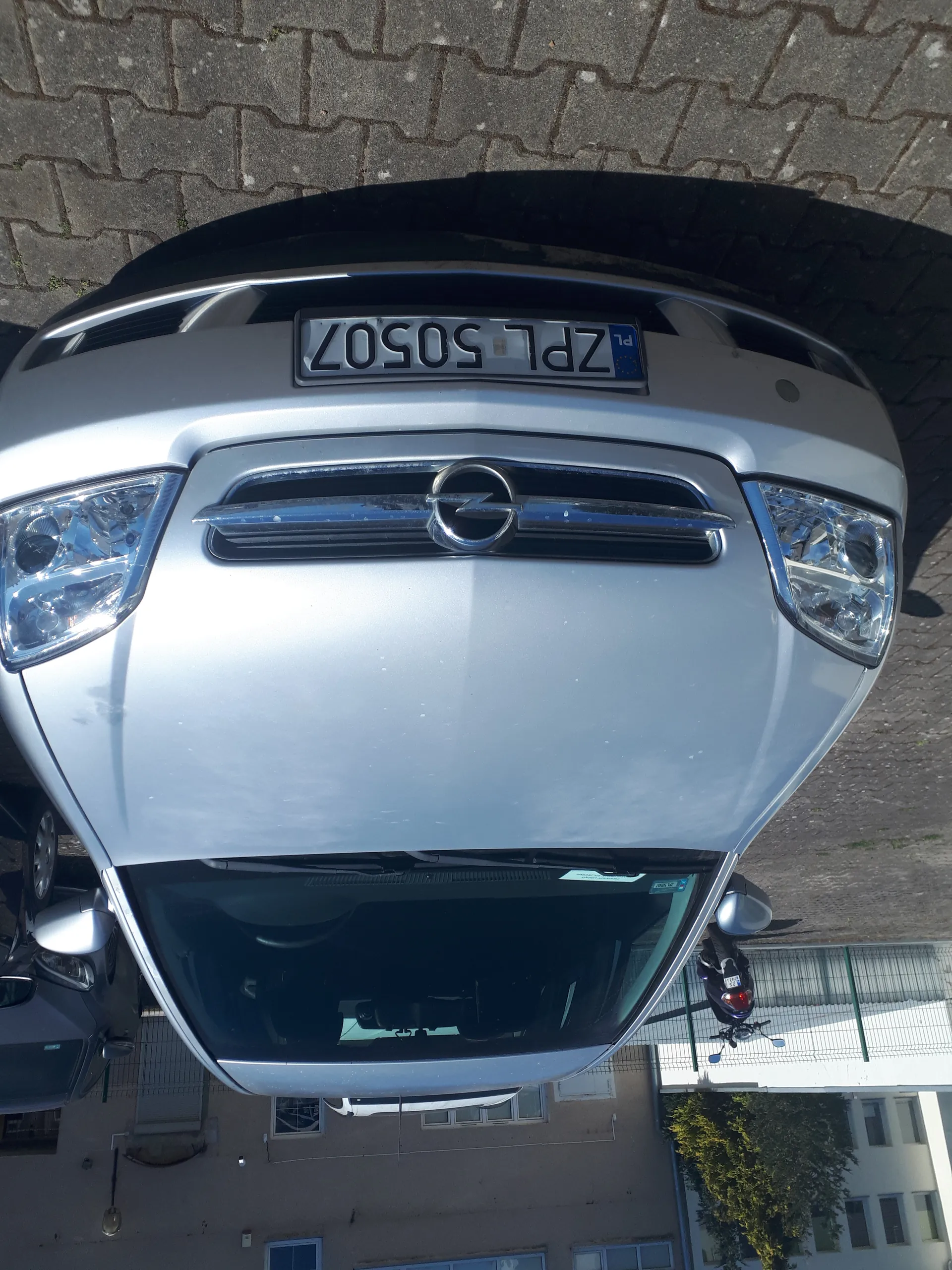 Srebrny samochód marki Opel z pionową tablicą rejestracyjną ZPL 50507, zaparkowany na brukowanej powierzchni. W tle widoczne są budynki, inny samochód oraz motocykl. Przednia szyba i reflektory są wyraźnie widoczne, a logo marki umieszczone jest na masce.