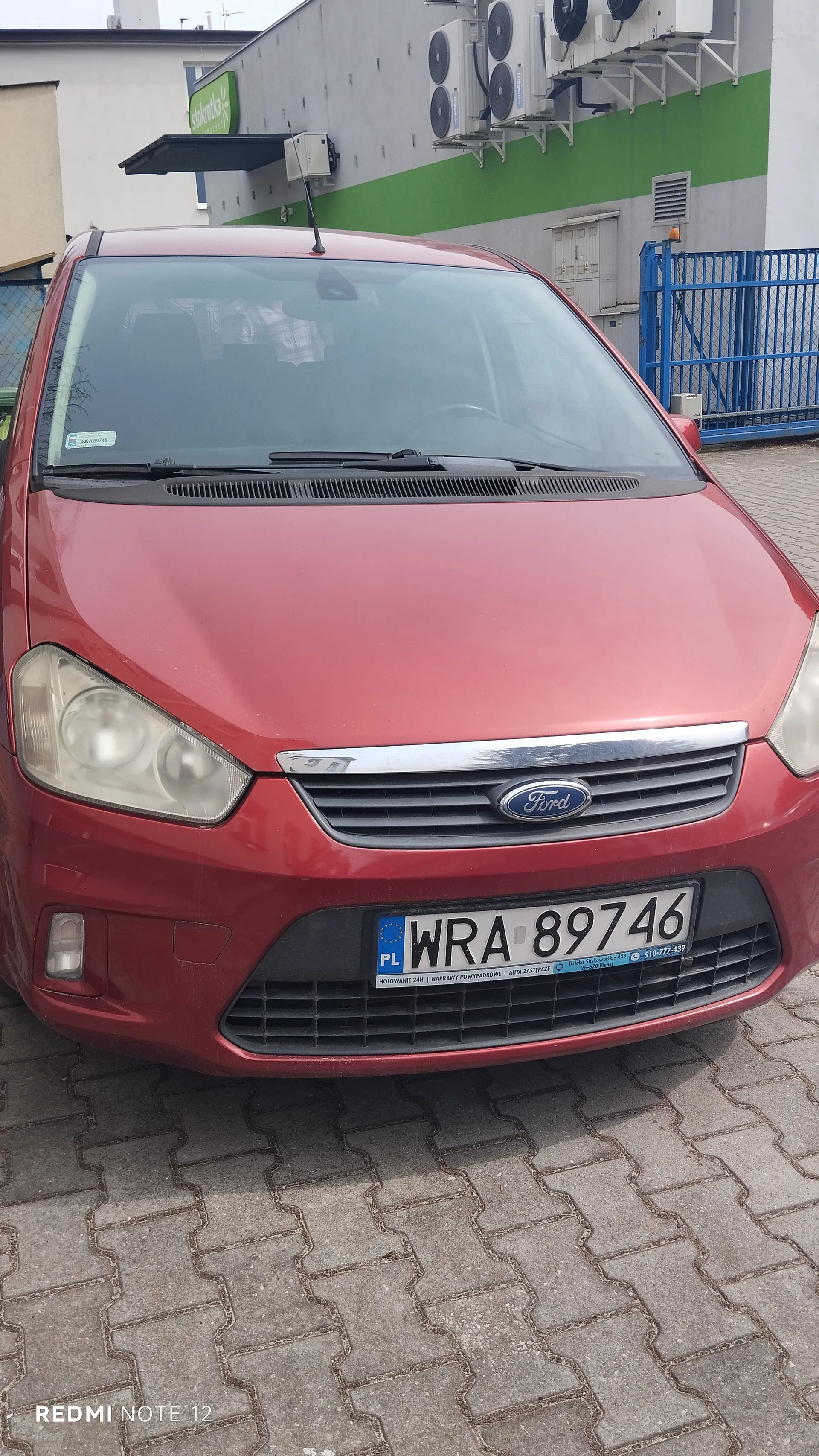 Czerwony samochód marki Ford z przodu, z widoczną tablicą rejestracyjną PL WRA 89746. Na tle budynek z zielono-białą elewacją, klimatyzatorami, niebieskim ogrodzeniem i zielonym szyldem sklepu stokrotka. Na dole zdjęcia widoczny napis REDMI NOTE 12. Samochód stoi na brukowanej powierzchni.