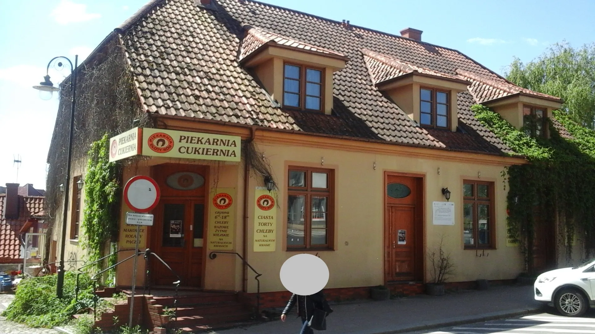 Parterowy budynek z poddaszem i brązowym, ceramicznym dachem. Na fasadzie znajduje się szyld z napisem „PIEKARNIA CUKIERNIA” oraz logo w kształcie okręgu. Obok wejścia widoczna jest biała tablica z ofertą: „ciasta, torty, chleby”. Budynek ma beżowe ściany, brązowe ramy okienne i drewniane drzwi. Po lewej stronie stoi latarnia uliczna, a po prawej części budynku pną się zielone rośliny. Przed budynkiem znajduje się biały samochód, a w centrum obrazu sylwetka osoby z zakrytym twarzą.