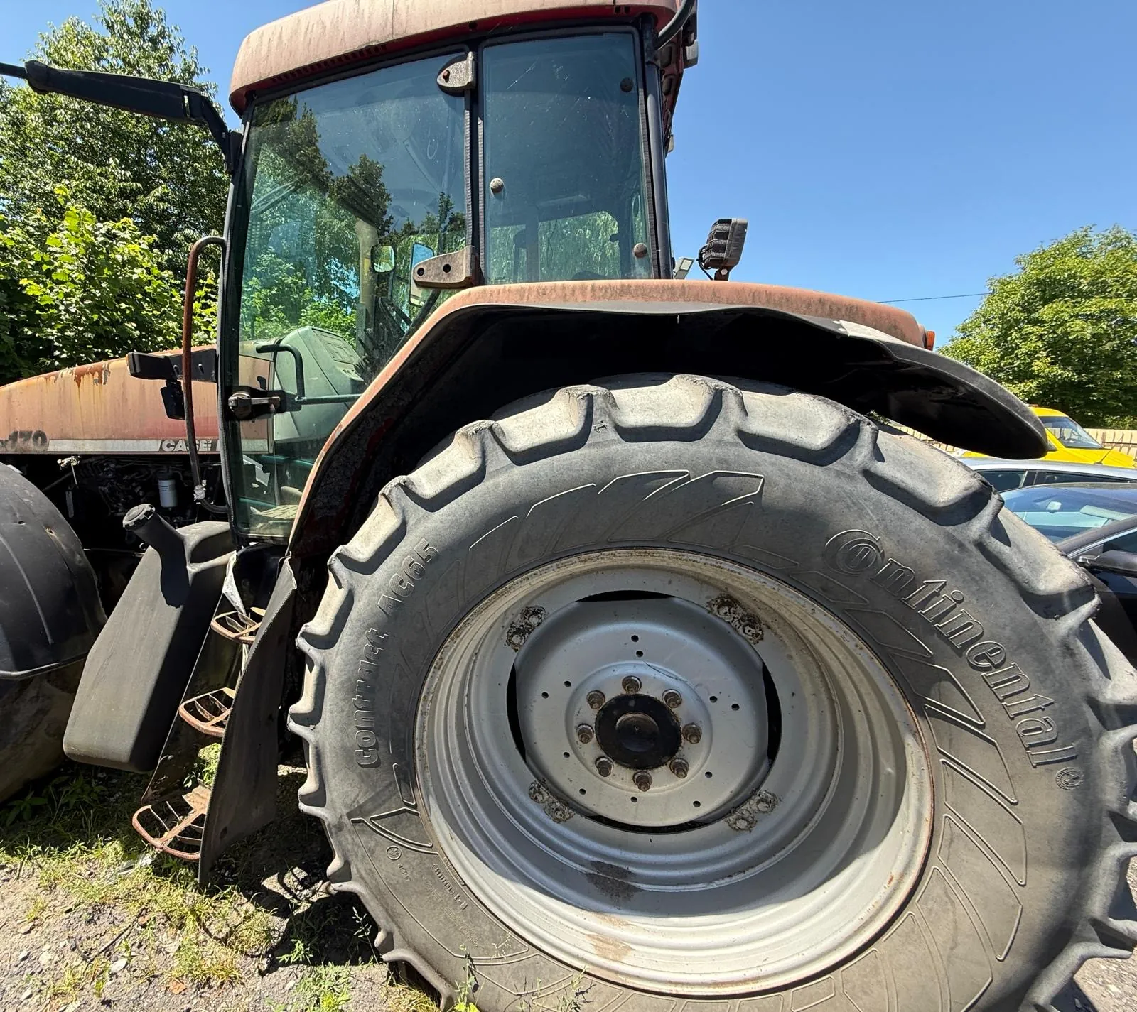 Zbliżenie na duże, tylne koło traktora marki Case IH. Opona jest szeroka, z wyraźnym bieżnikiem i napisem 'Continental' oraz 'ComfortForce'. Felga jest metalowa, w kolorze srebrnym. W tle widoczna jest kabina traktora, zielone drzewa oraz fragmenty zaparkowanych samochodów pod bezchmurnym, niebieskim niebem.