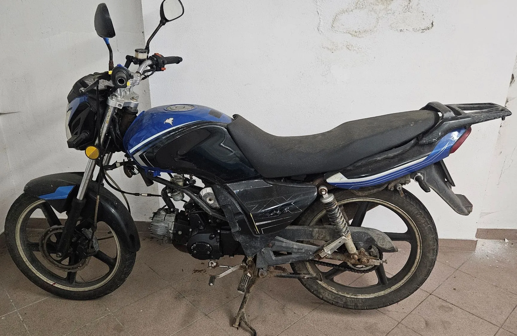 Niebiesko-czarny motocykl zaparkowany w pomieszczeniu na jasnym tle. Pojazd ma czarne felgi, czarne siedzenie i widoczne elementy silnika oraz układu wydechowego. Motocykl stoi na bocznej stopce.
