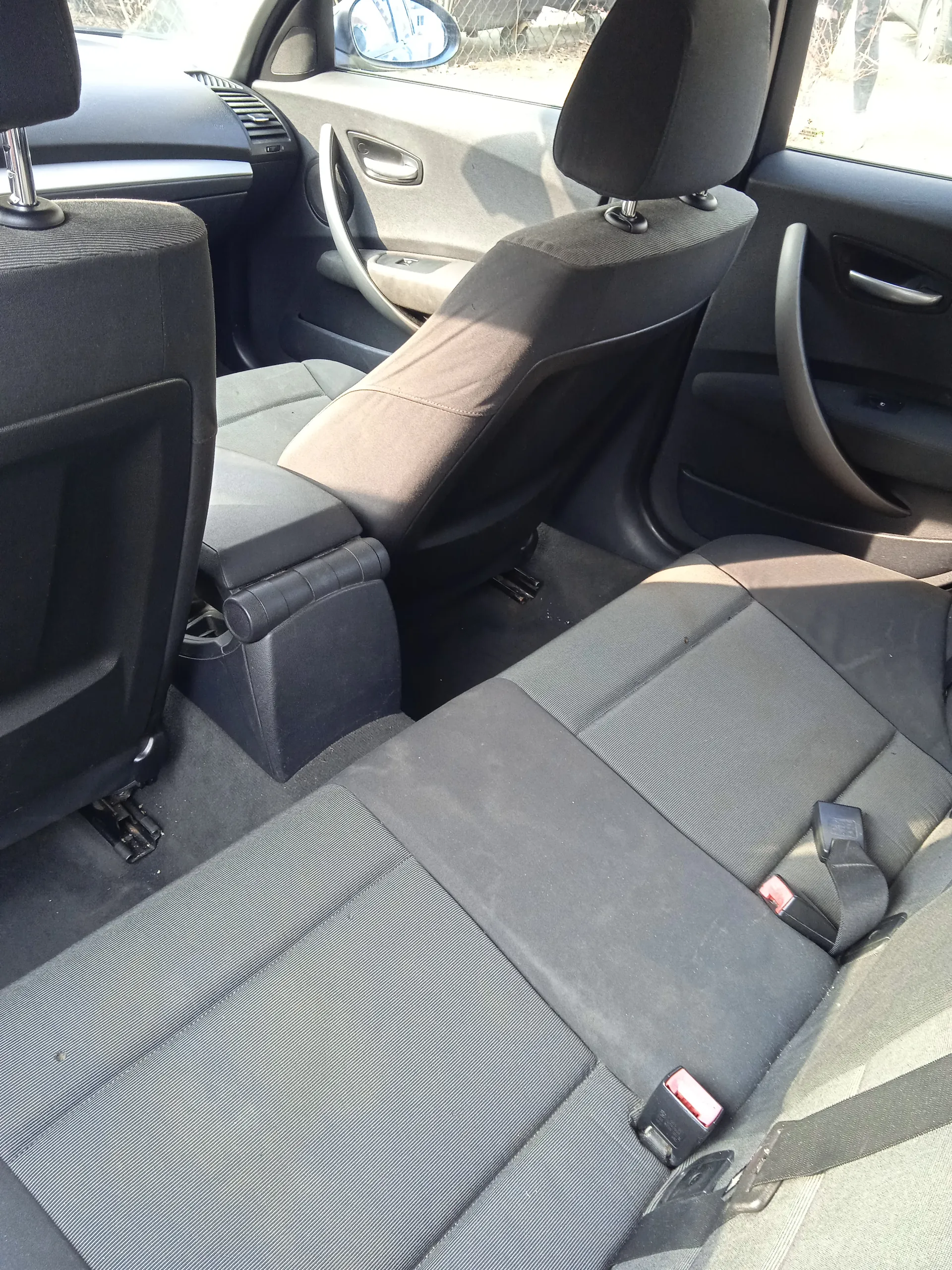 Wnętrze samochodu widziane z tyłu. Widoczne są czarne tkaninowe siedzenia przednie i tylne, zagłówki, pasy bezpieczeństwa z czerwonymi i czarnymi zapięciami. W tle widać kierownicę, deski rozdzielczą, drzwi pasażera z uchwytem i lusterkiem bocznym. Przez okna widoczny jest zewnętrzny krajobraz z ogrodzeniem i drzewami. Brak czytelnego tekstu.